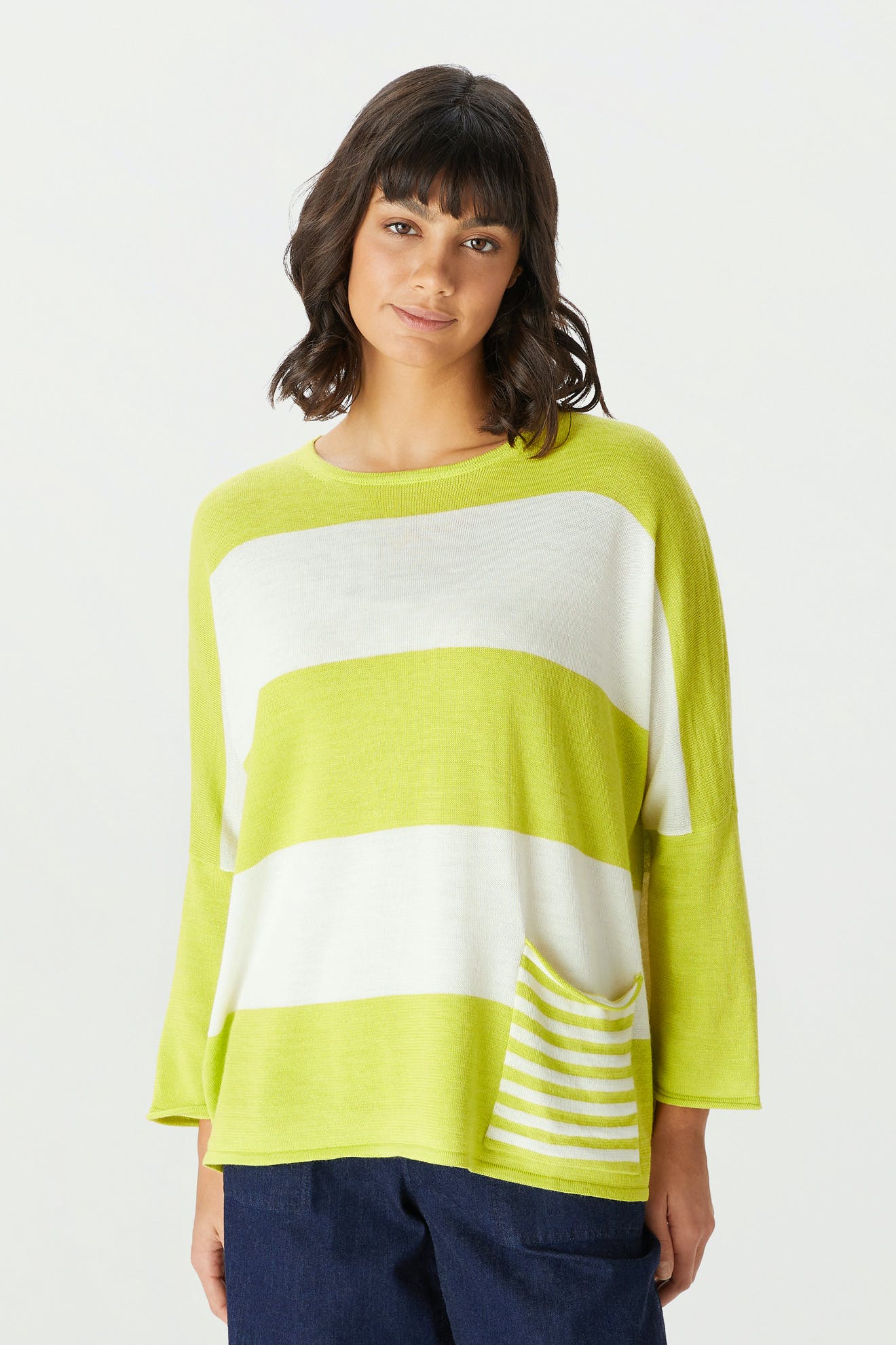 A woman wearing the Merino Blend Stripe Sweater—a loose-fitting, long-sleeved sweater featuring wide horizontal lime green and white stripes and a matching striped pocket—paired with dark pants, stands against a plain background, making it the perfect transitional weather sweater.