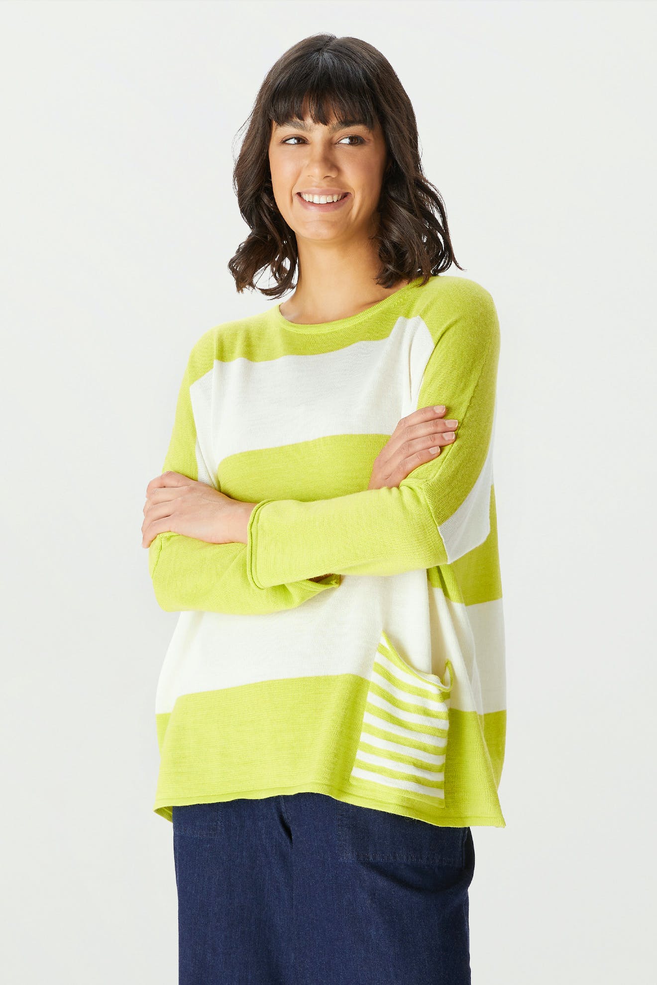 A woman with shoulder-length dark hair stands with her arms crossed and smiles against a plain light background, wearing the Merino Blend Stripe Sweater in white and light green along with dark pants.