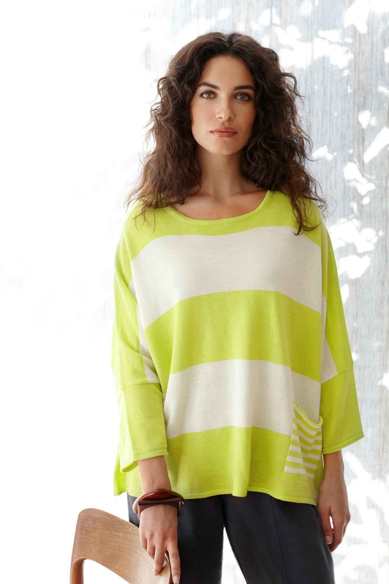 A woman with curly hair stands indoors by a chair, sunlight filtering through the window behind her, wearing the Merino Blend Stripe Sweater in loose-fitting lime green and white stripes—a perfect choice for anyone seeking a cozy sweater for transitional weather.