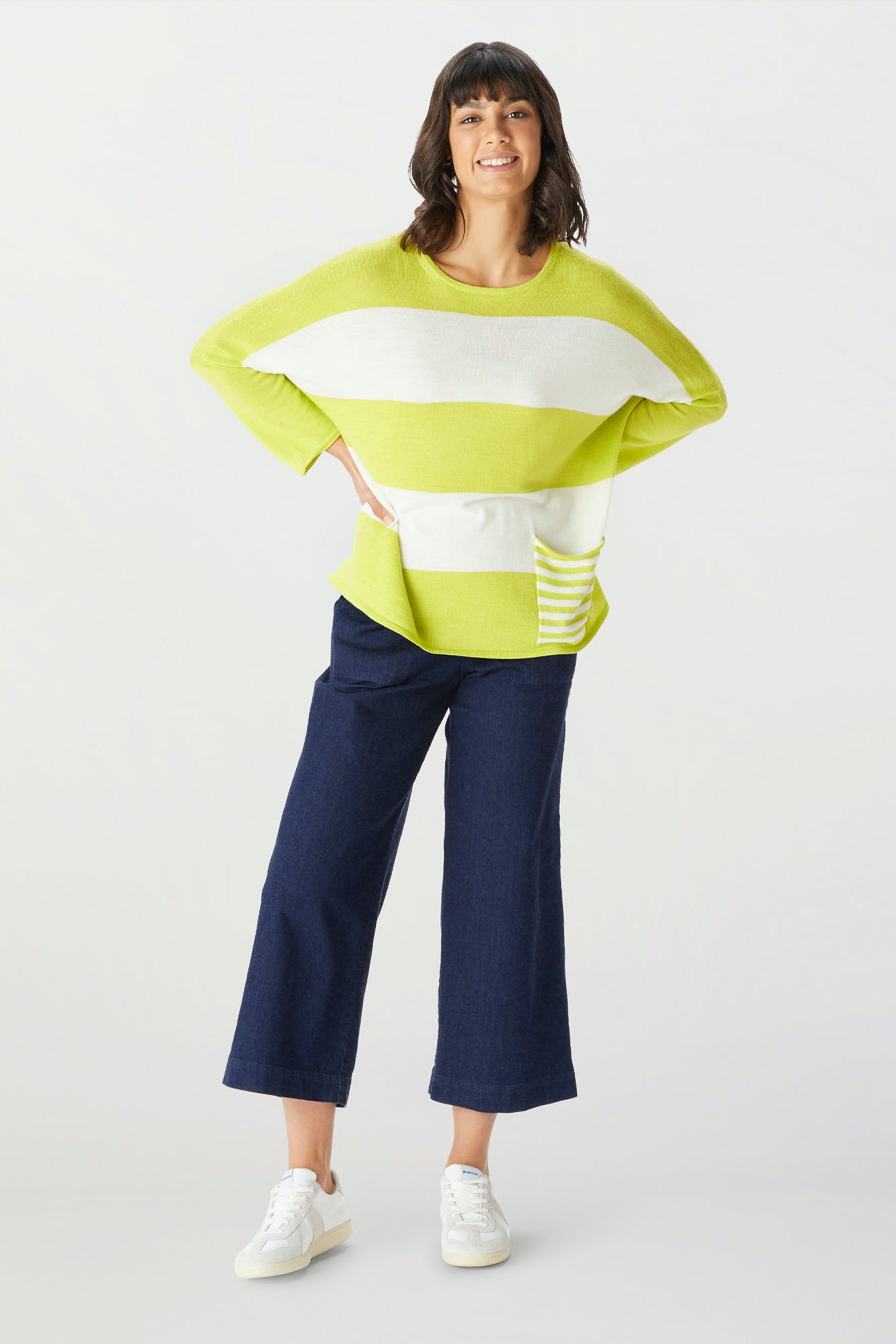 A woman stands and smiles in the Merino Blend Stripe Sweater—lime green and white stripes make it perfect as a transitional weather sweater—paired with wide-leg dark blue jeans and white sneakers against a plain light background.