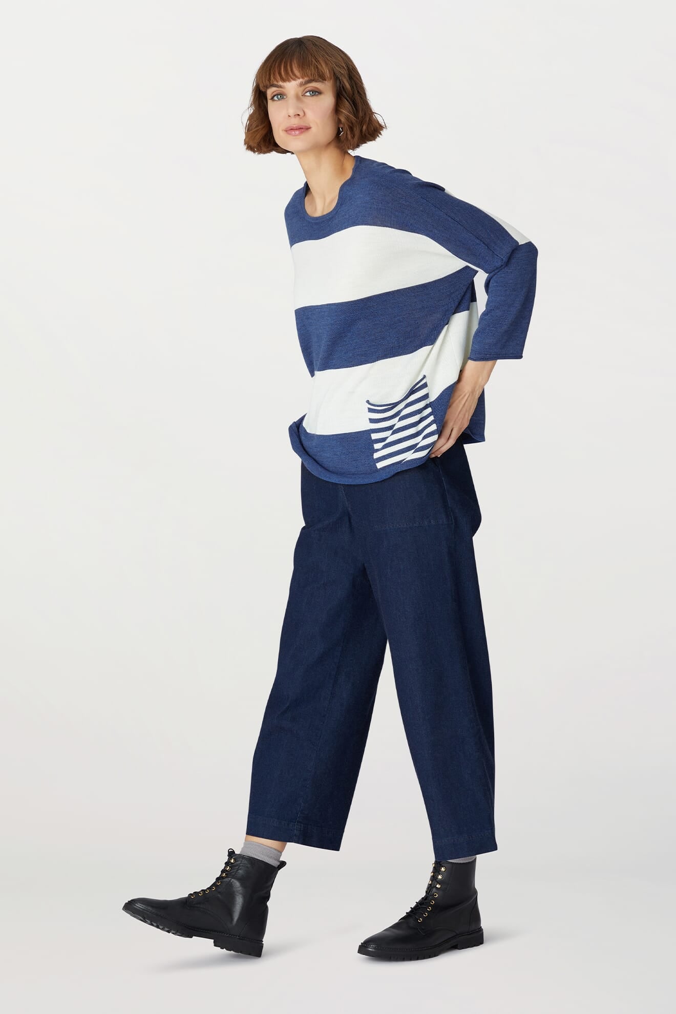 A woman wears the Merino Blend Stripe Sweater, featuring blue and white stripes with pocket detail, paired with wide-leg dark blue pants and black boots. She stands against a plain white background—an ideal look for transitional weather.