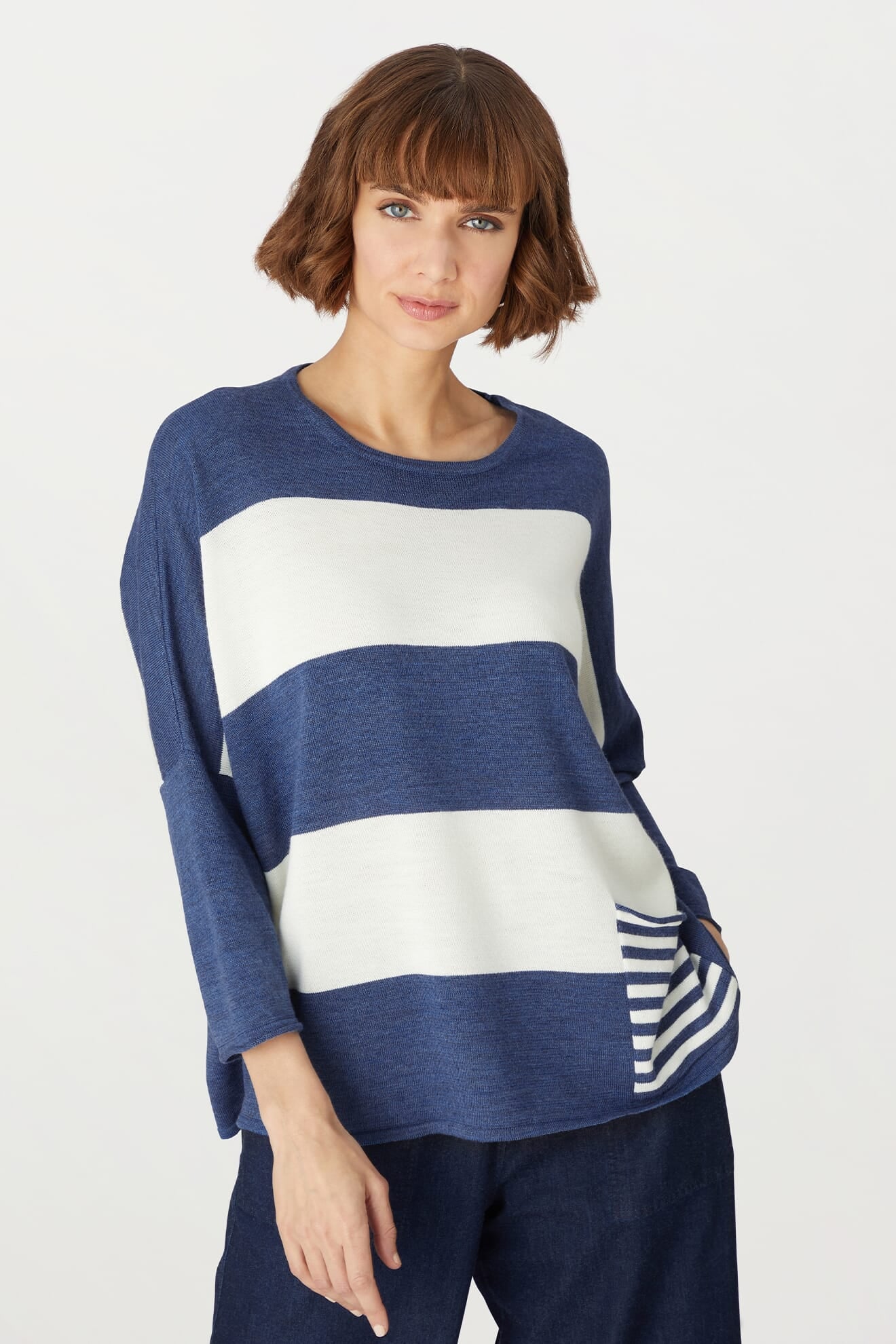 A woman stands against a plain white background, wearing the Merino Blend Stripe Sweater—a blue and white color-blocked long-sleeve top featuring a striped pocket detail—paired with dark pants. This is the perfect transitional weather sweater, combining both style and comfort.