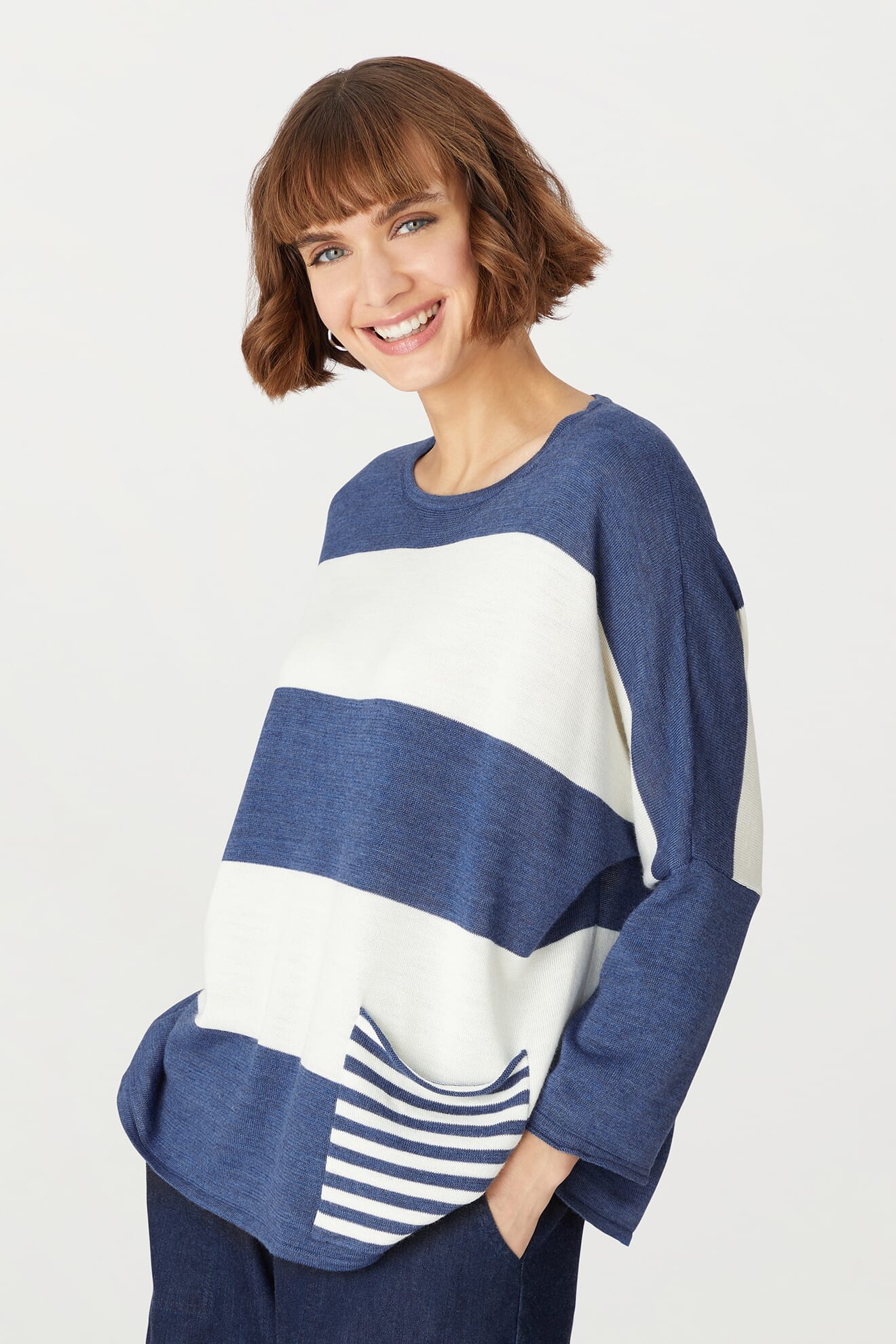 Wearing the Merino Blend Stripe Sweater in blue and white stripes, featuring a convenient front pocket, this person pairs it with dark pants while smiling against a plain white background. The Merino Blend Stripe Sweater is a cozy choice for transitional weather, perfect for adding both style and comfort to any wardrobe.