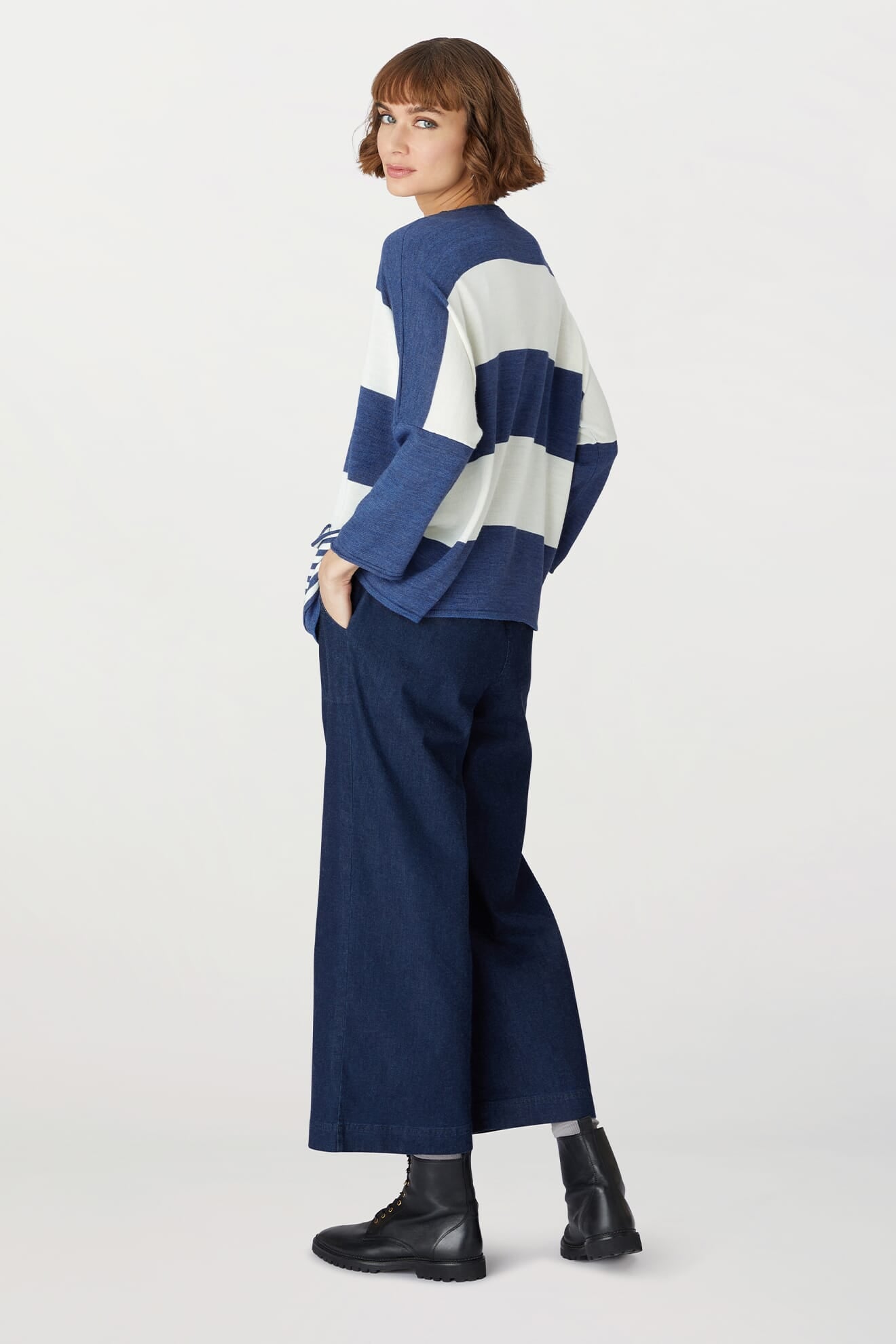 A woman stands sideways against a plain white background, wearing the Merino Blend Stripe Sweater—perfect for transitional weather—paired with wide-leg dark blue jeans and black boots.