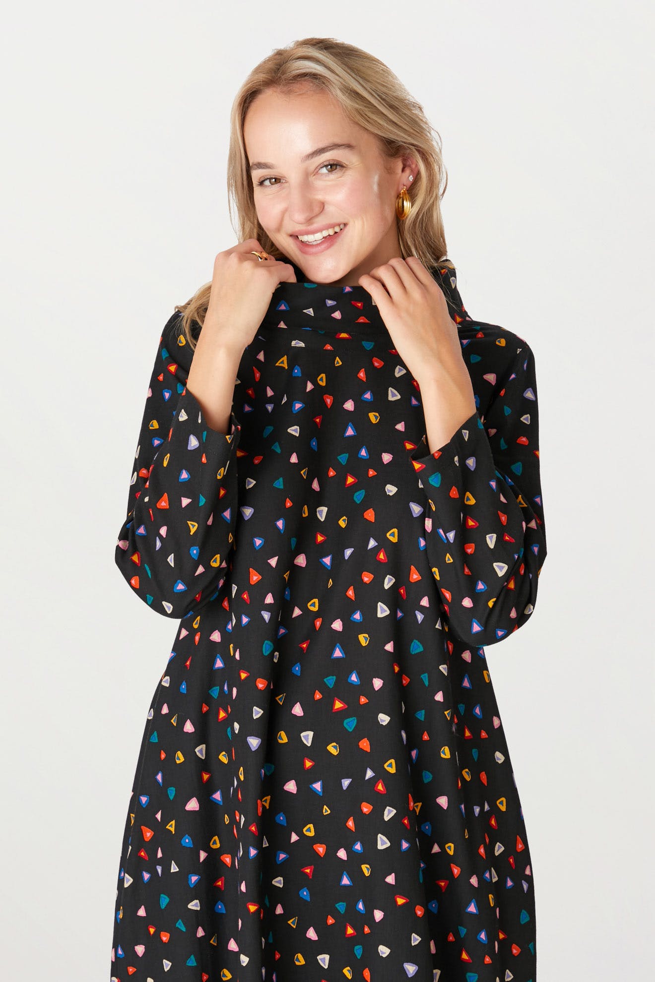 Woman in Little Graphic Triangle Dress smiling at the camera, with hands touching her neck, standing against a white background.