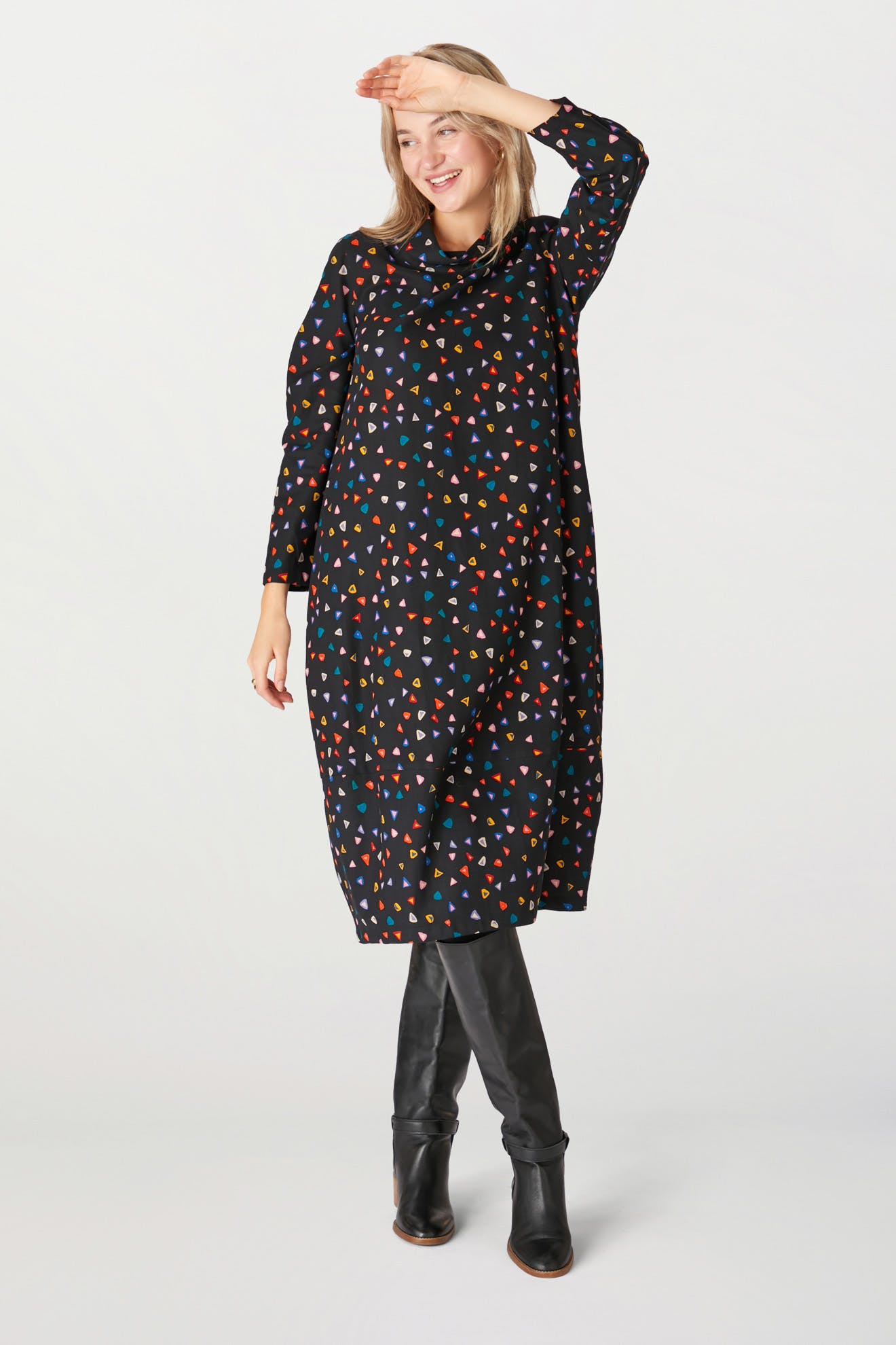 A woman in a Little Graphic Triangle Dress with colorful geometric patterns and knee-high boots, smiling and posing with one hand on her head.