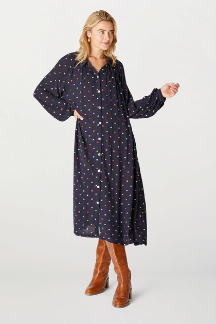 Woman standing in a Small Square Embroidered Dress with multicolored polka dots, paired with brown leather boots, against a light background.