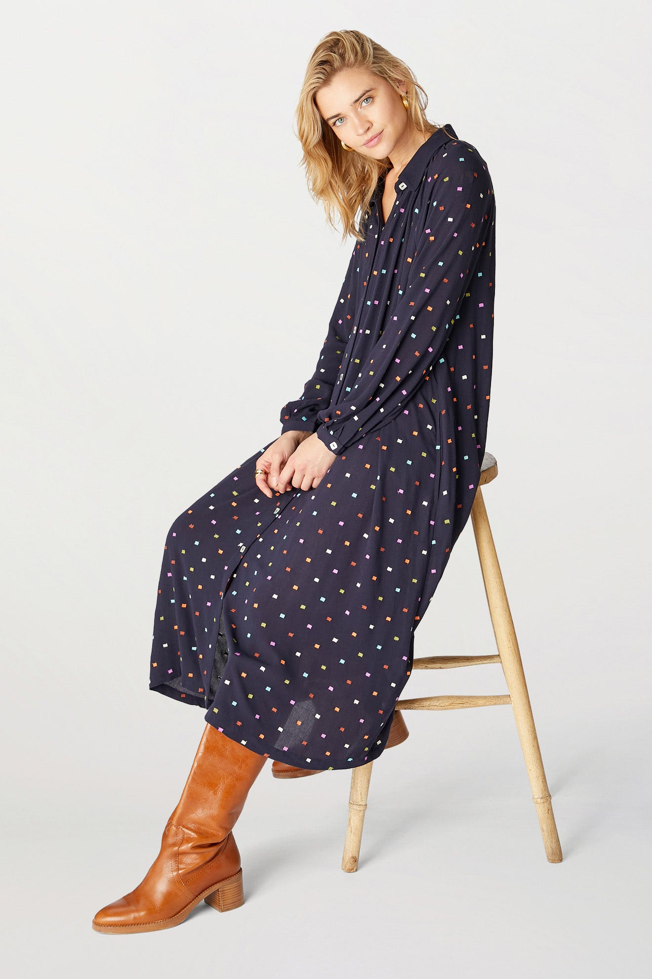 Woman in a Small Square Embroidered Dress sitting on a wooden stool, wearing brown boots, against a white background.