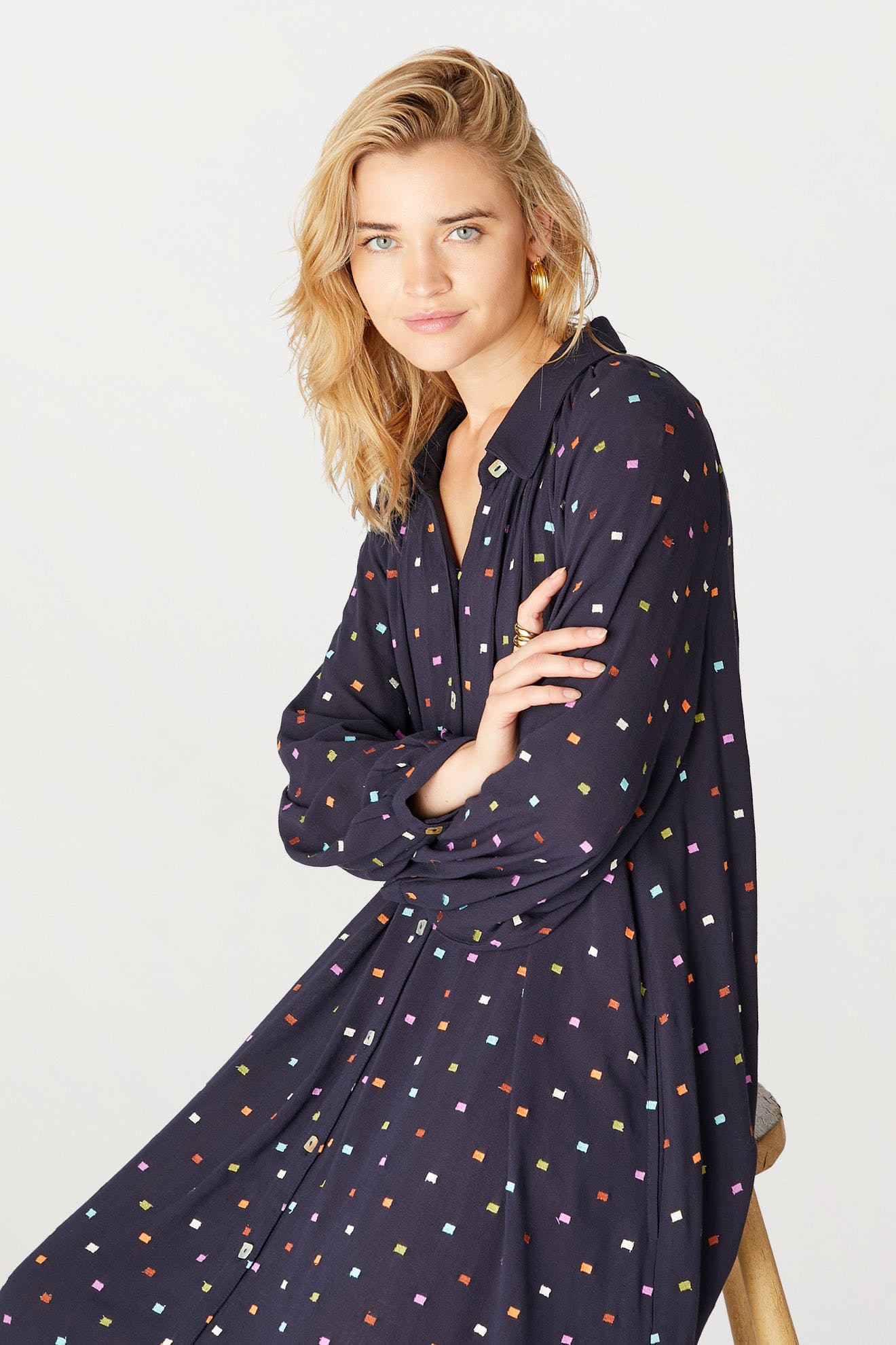 Woman with blonde hair posing in a Small Square Embroidered Dress with colorful polka dots, leaning on a wooden stool, against a white background.