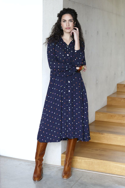 Woman in a Small Square Embroidered Dress and brown boots standing beside wooden stairs, touching her face thoughtfully.