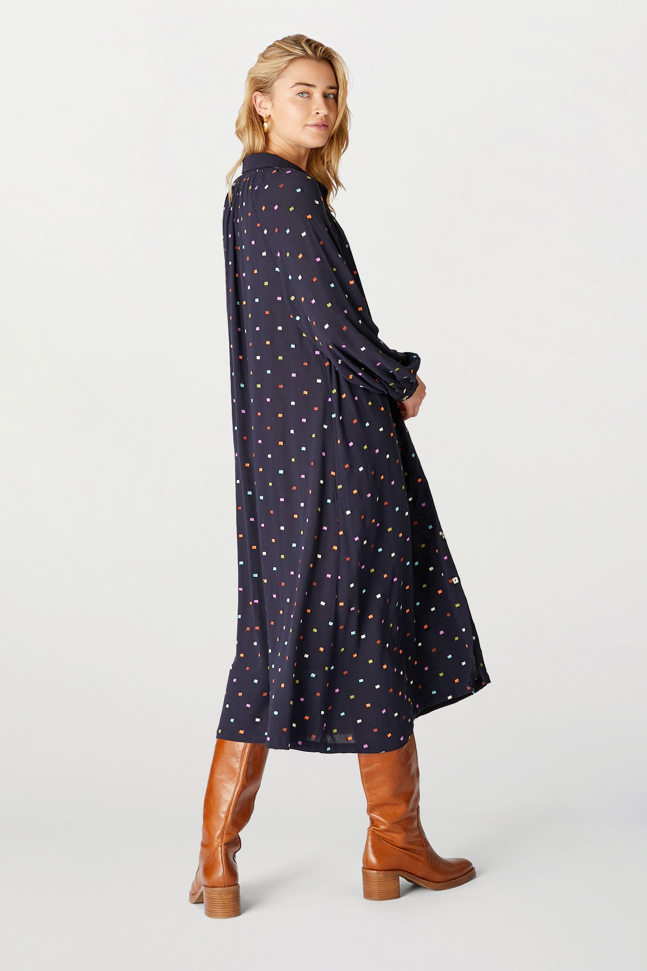 Woman in a navy blue Small Square Embroidered Dress with colorful polka dots and brown knee-high boots, standing sideways and looking over her shoulder.