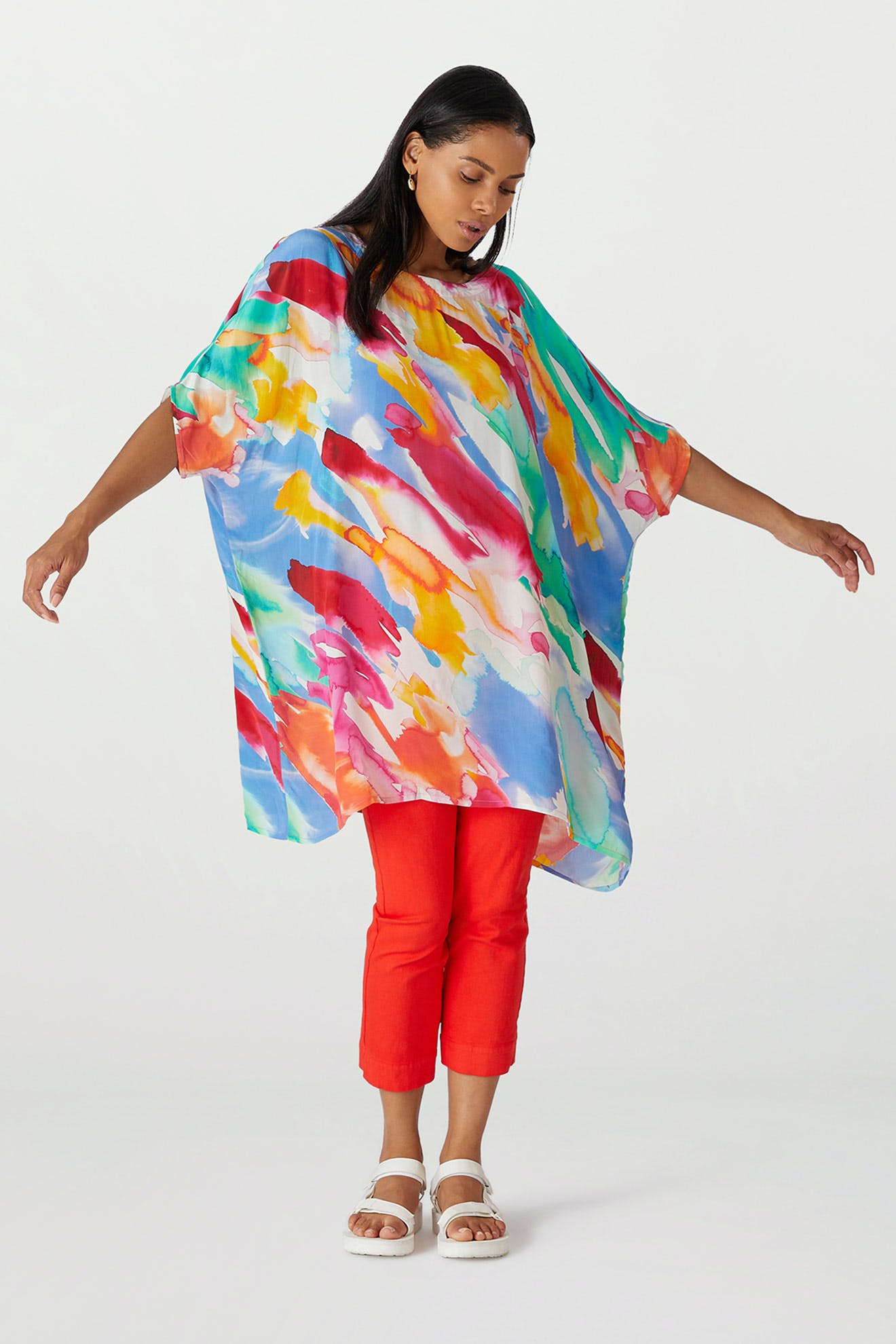 A woman wearing a Rainbow Splash Viscose Tunic over red trousers and white sandals poses with arms outstretched against a plain background.