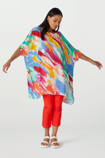 A woman wearing a Rainbow Splash Viscose Tunic over red trousers and white sandals poses with arms outstretched against a plain background.