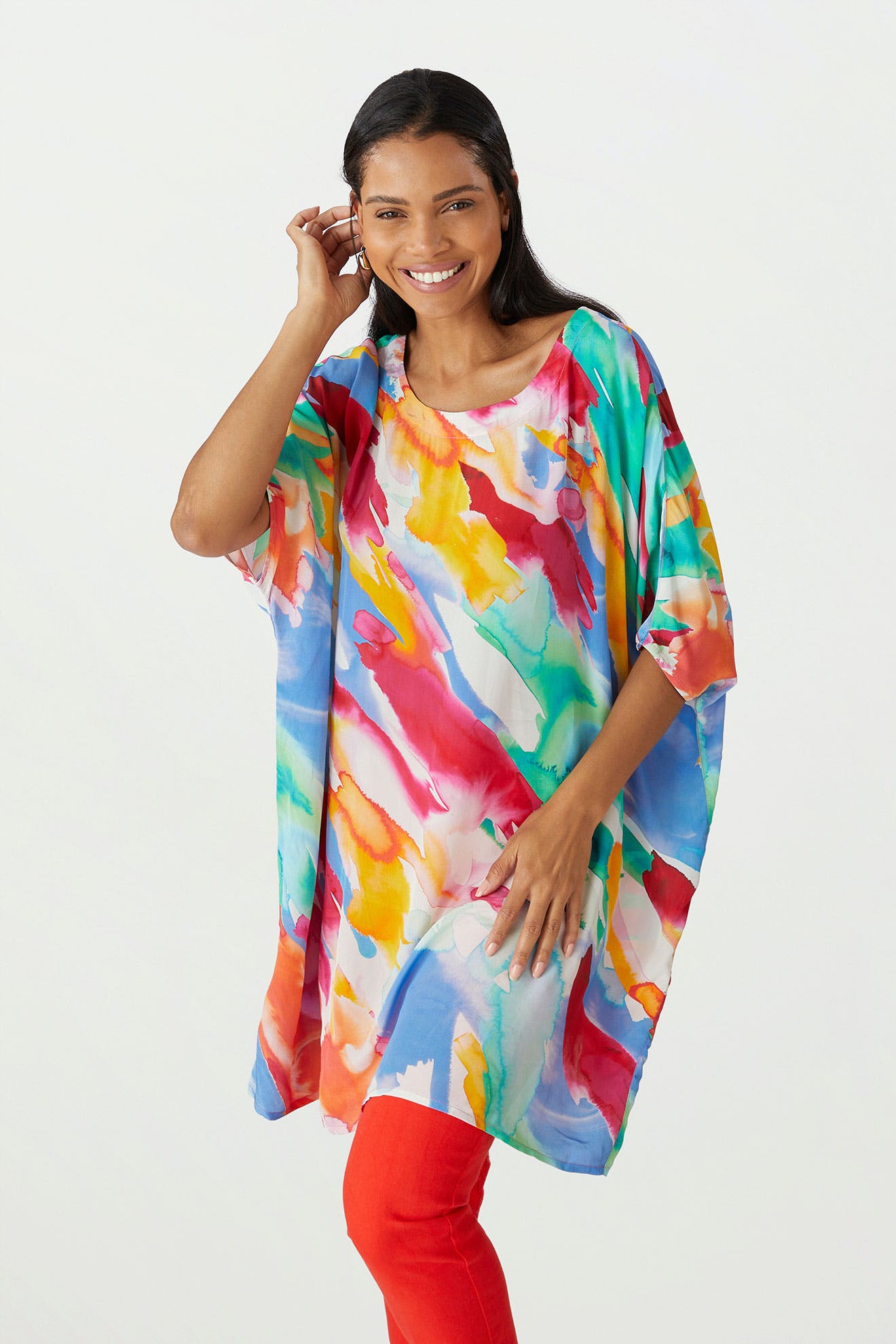 Woman smiling, standing in a studio, wearing a Rainbow Splash Viscose Tunic with painterly motifs and red pants. She is touching her hair with one hand. White background.