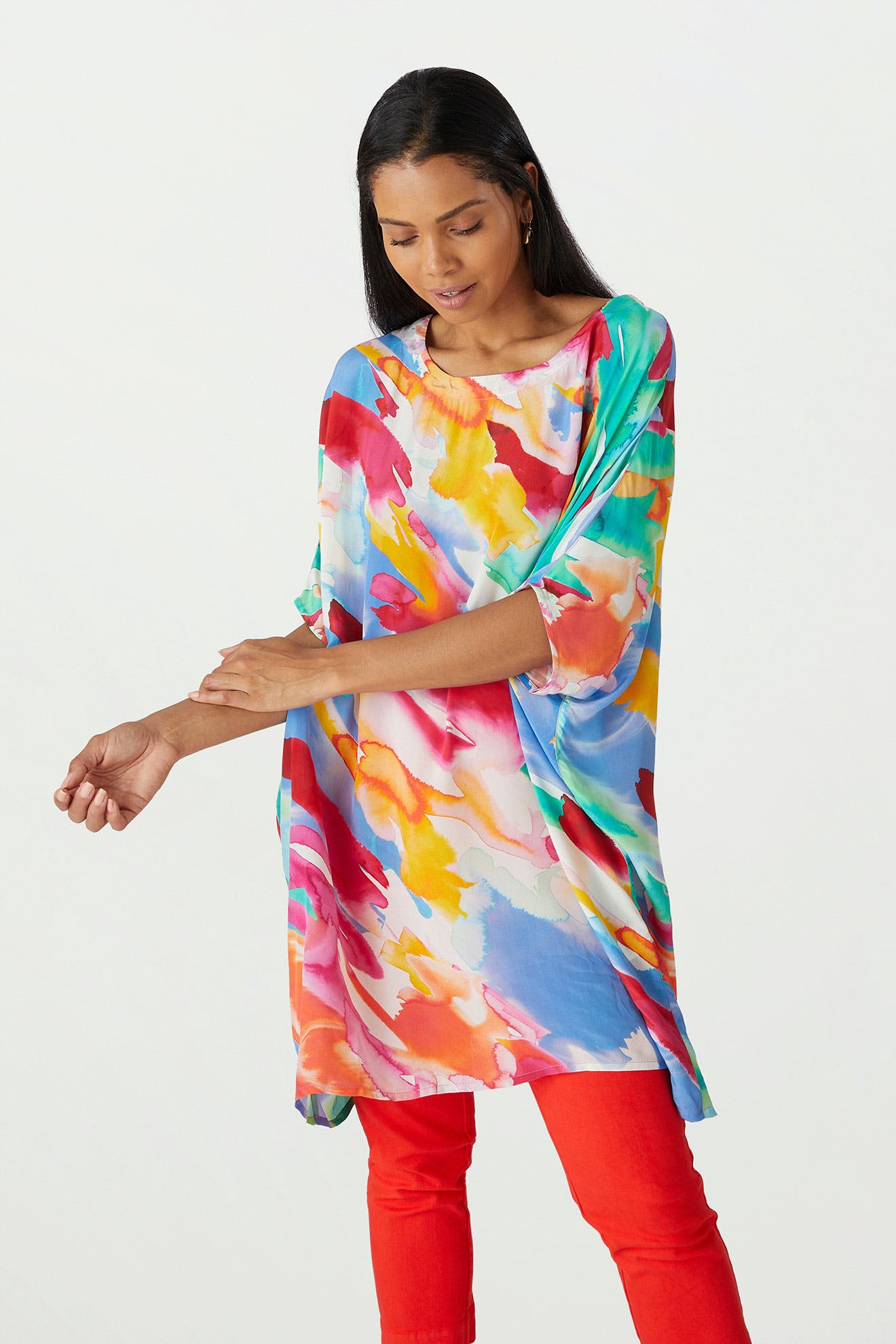 A woman in a Rainbow Splash Viscose Tunic with painterly motifs and red pants checking her watch.