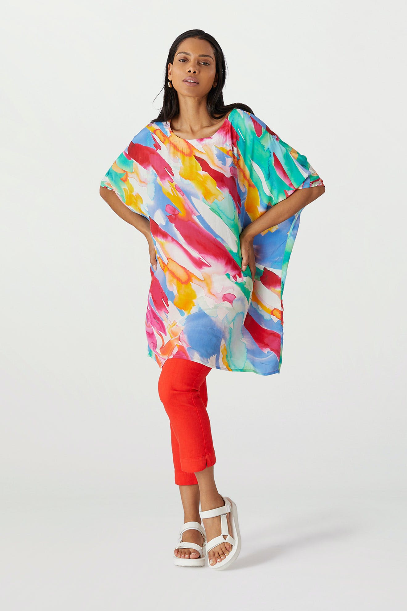 Woman standing in a studio, wearing a Rainbow Splash Viscose tunic and red trousers, paired with white sandals.