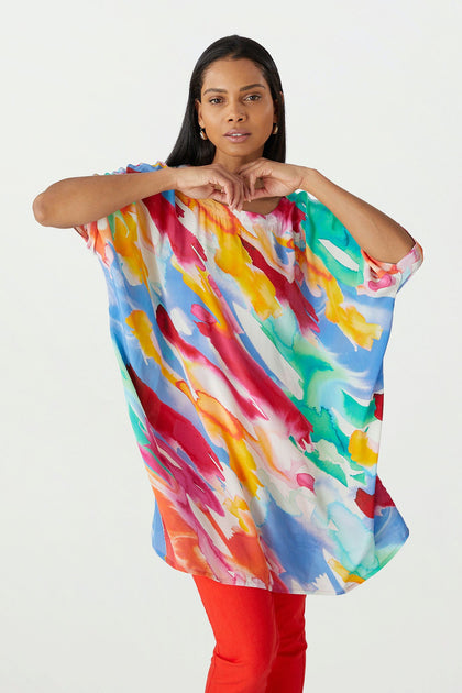 A woman standing in a studio, wearing the Rainbow Splash Viscose Tunic and red pants, makes a heart shape with her hands.