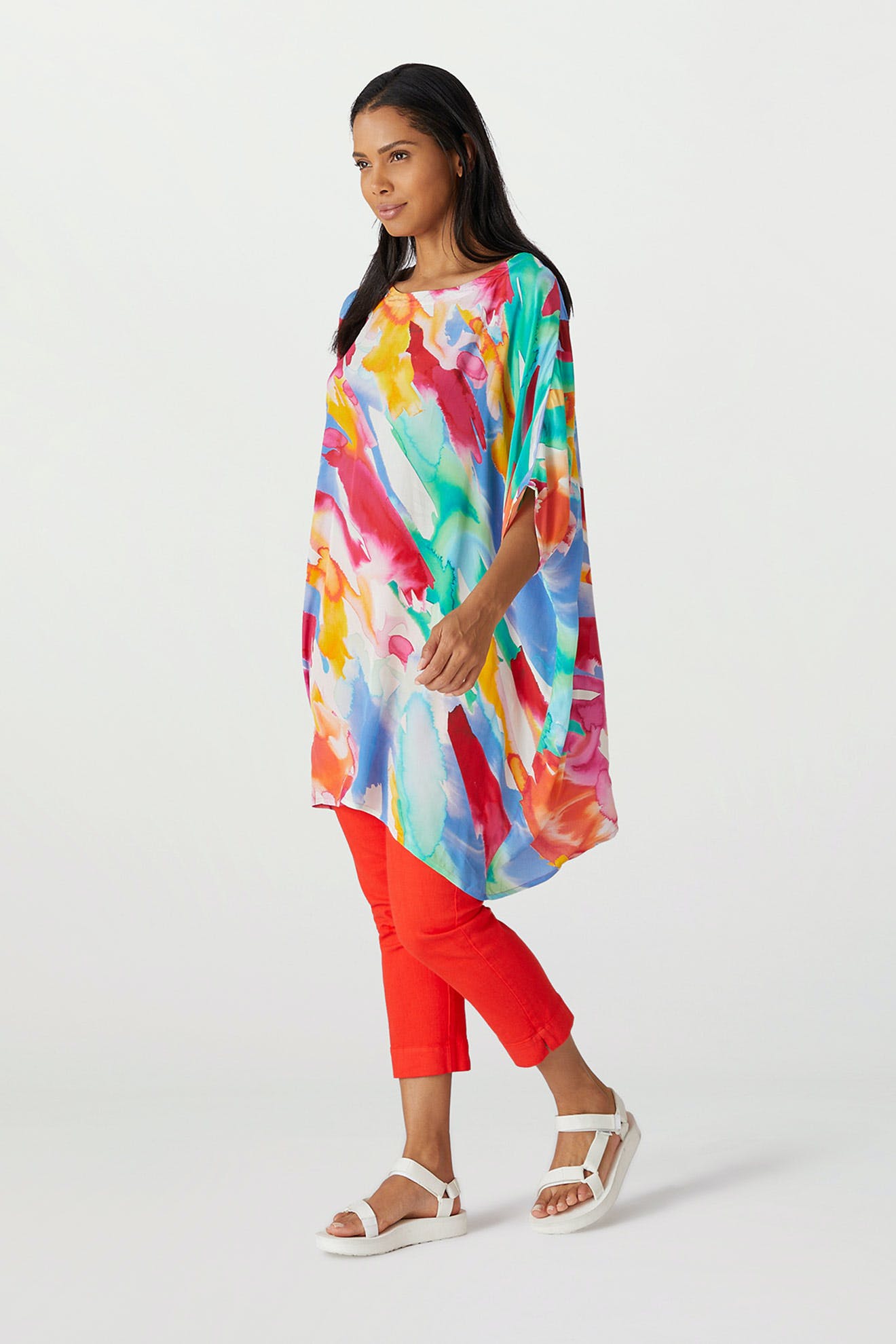 Woman posing in a Rainbow Splash Viscose Tunic with a painterly motif and red pants, walking to the side against a white background.