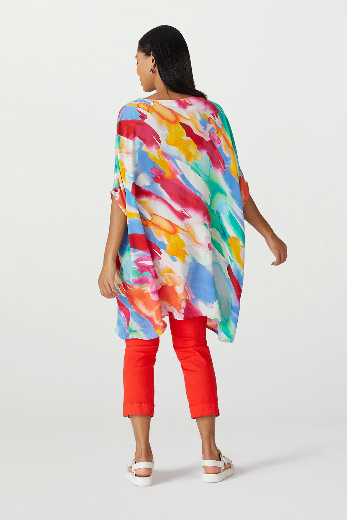 A woman stands facing away, wearing a Rainbow Splash Viscose Tunic and red capri pants, paired with white sandals.