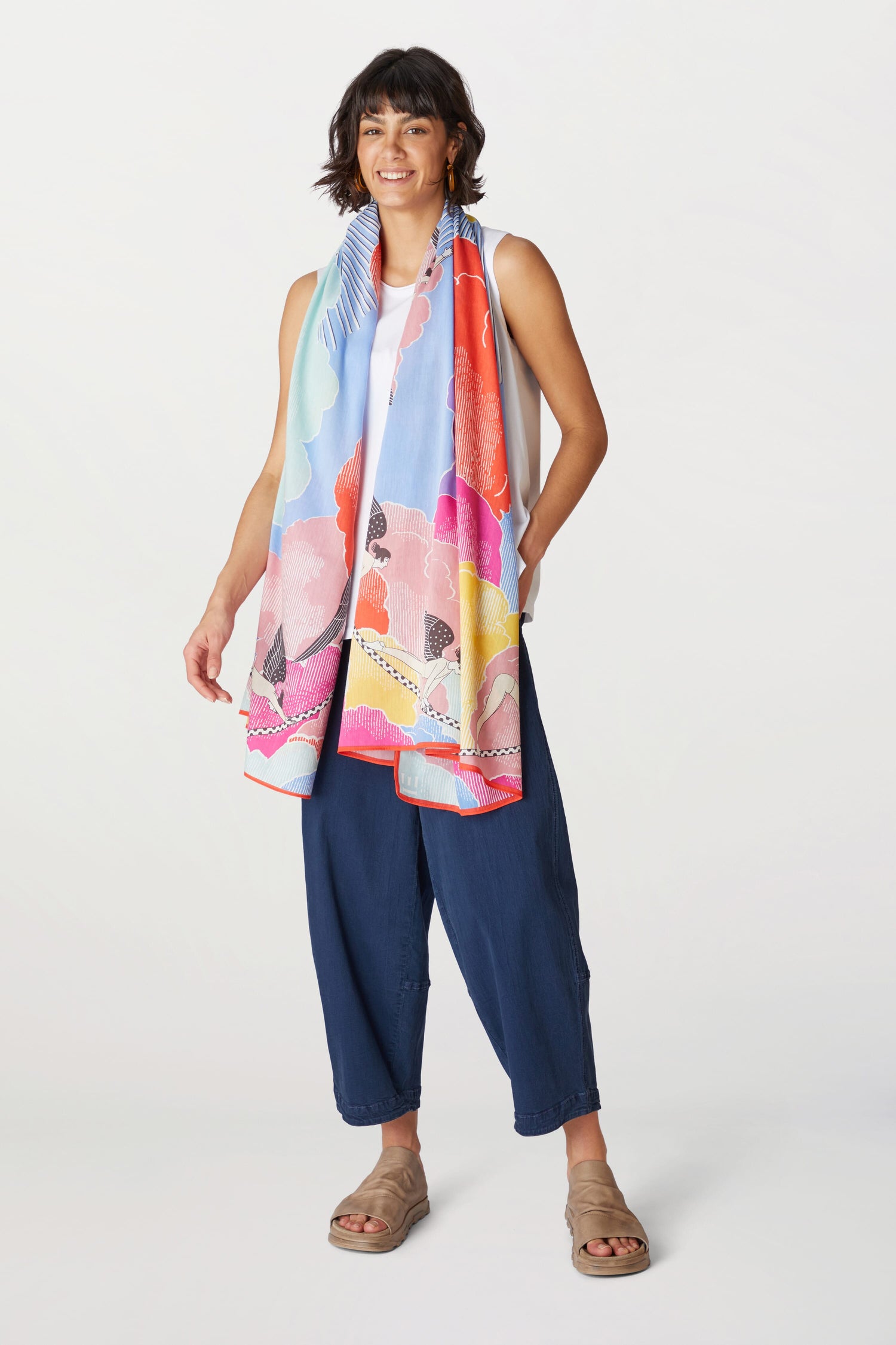 A woman stands smiling, wearing a Yoga Flow Scarf with a unique sunrise-inspired print, a white top, navy blue cropped pants, and beige sandals against a white background.