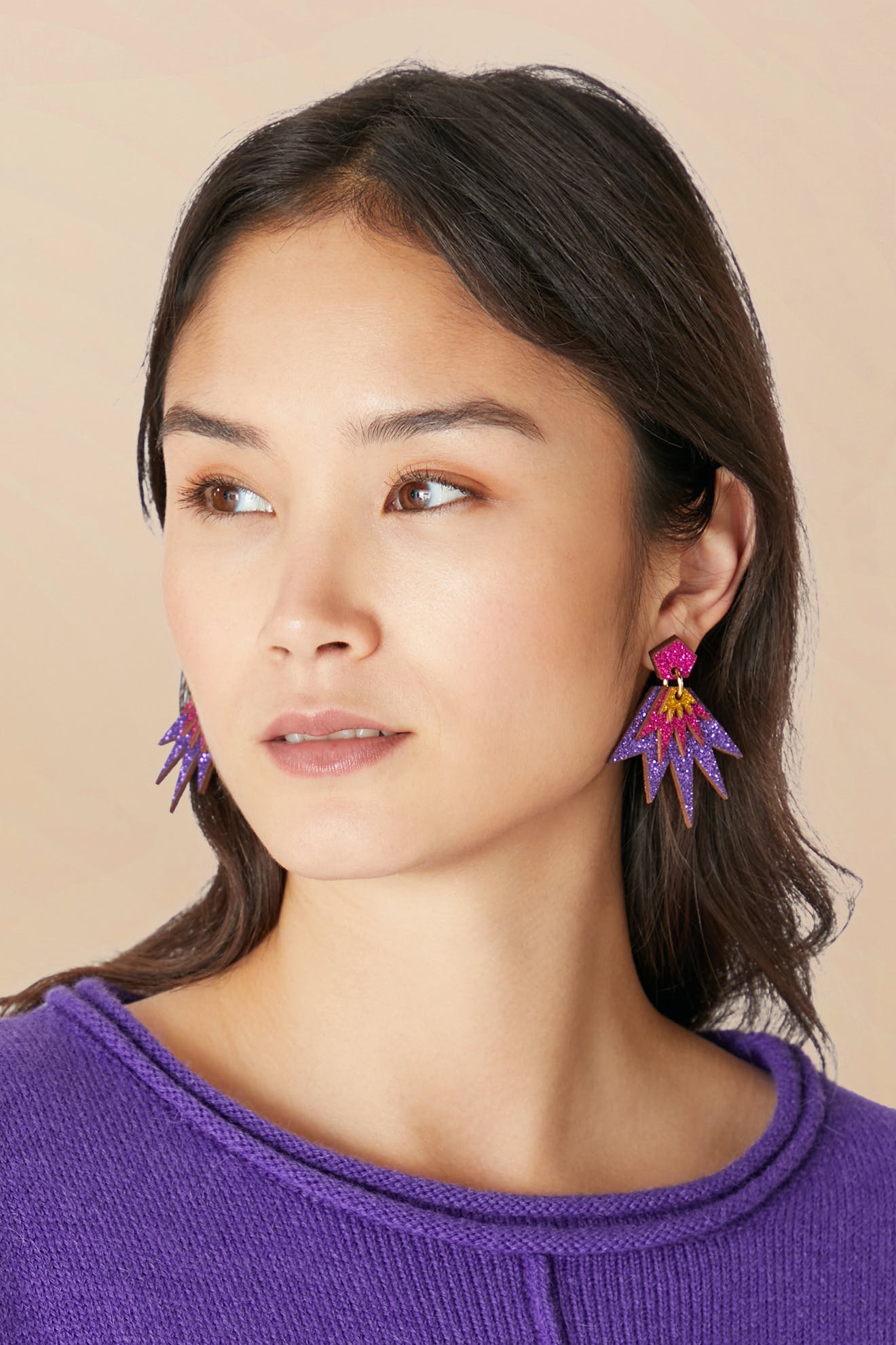 A woman with dark hair wears vibrant Sparkle Drop Earrings, shaped like stars in purple and pink. She is also dressed in a purple knit top and gazes slightly to the side against a neutral background.