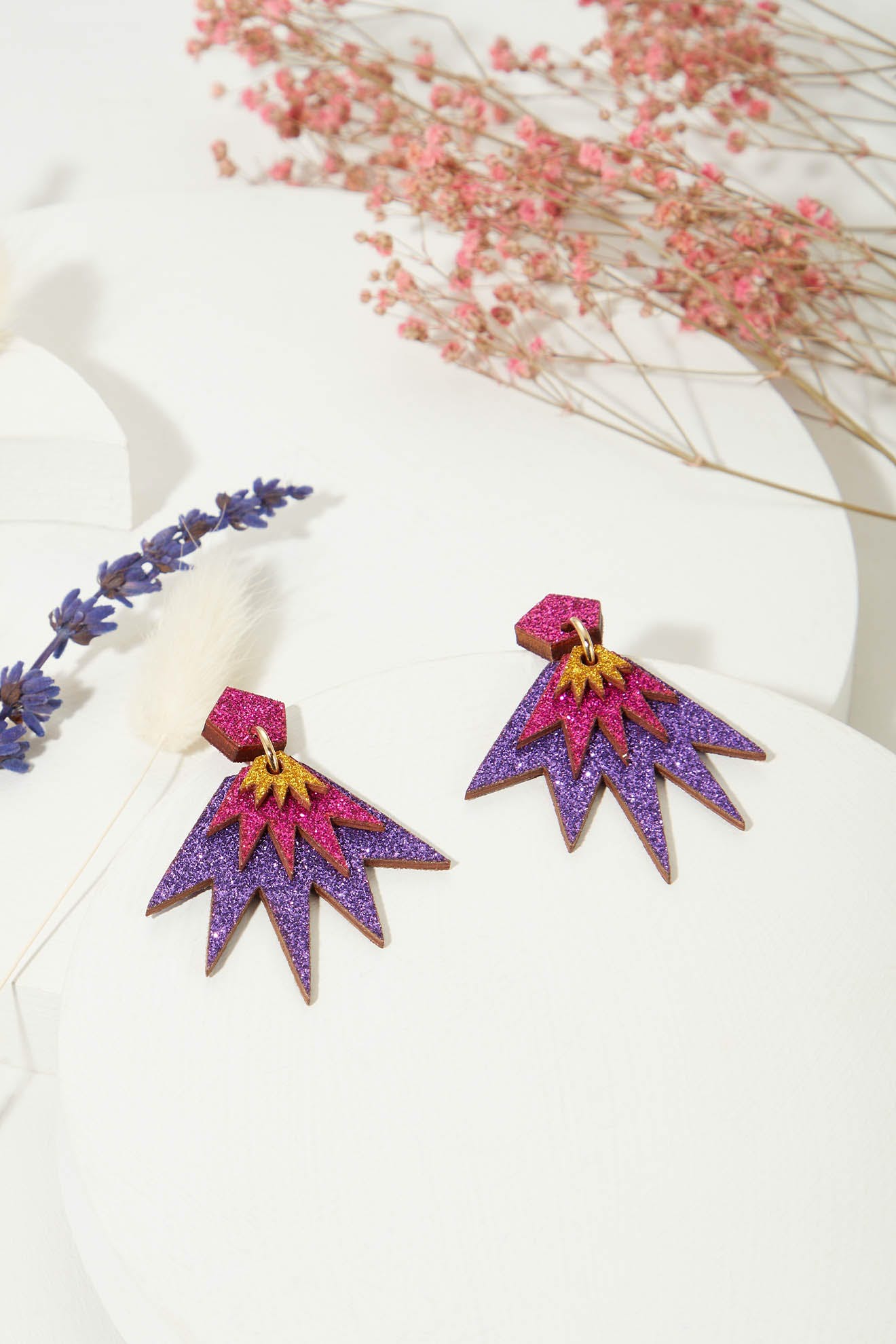 The Sparkle Drop Earrings, featuring a pair of colorful, starburst-shaped lightweight drops adorned with non-shedding purple and pink glitter, are displayed on a white surface surrounded by sprigs of lavender and dried pink flowers.