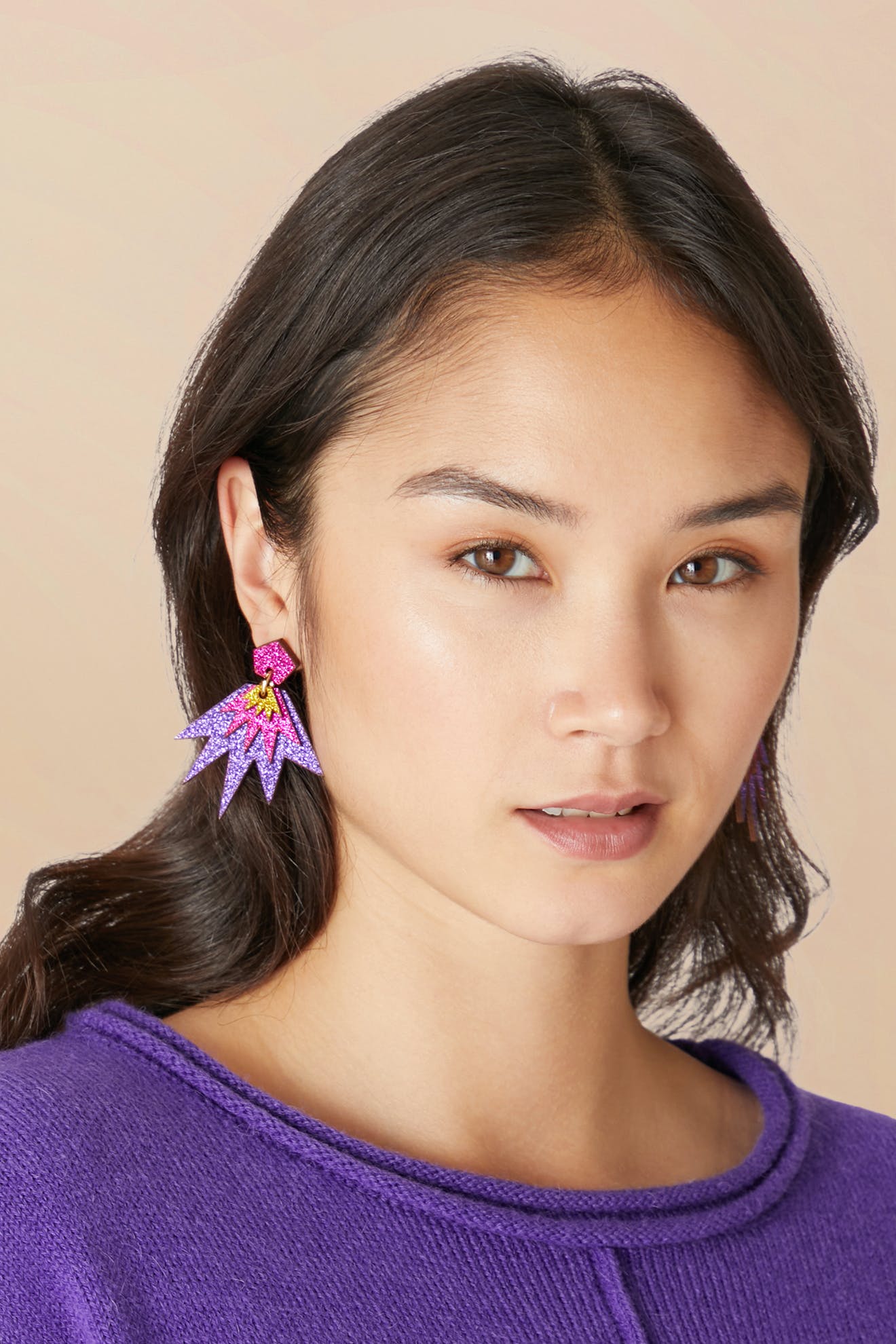 A woman with shoulder-length hair wears Sparkle Drop Earrings in purple and pink geometric designs, crafted from eco-friendly wood, paired with a purple top, posing against a neutral background.