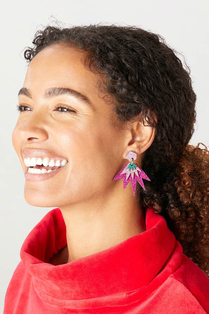 A person wearing a red top smiles, showcasing large, colorful Sparkle Drop Earrings shaped like starbursts.