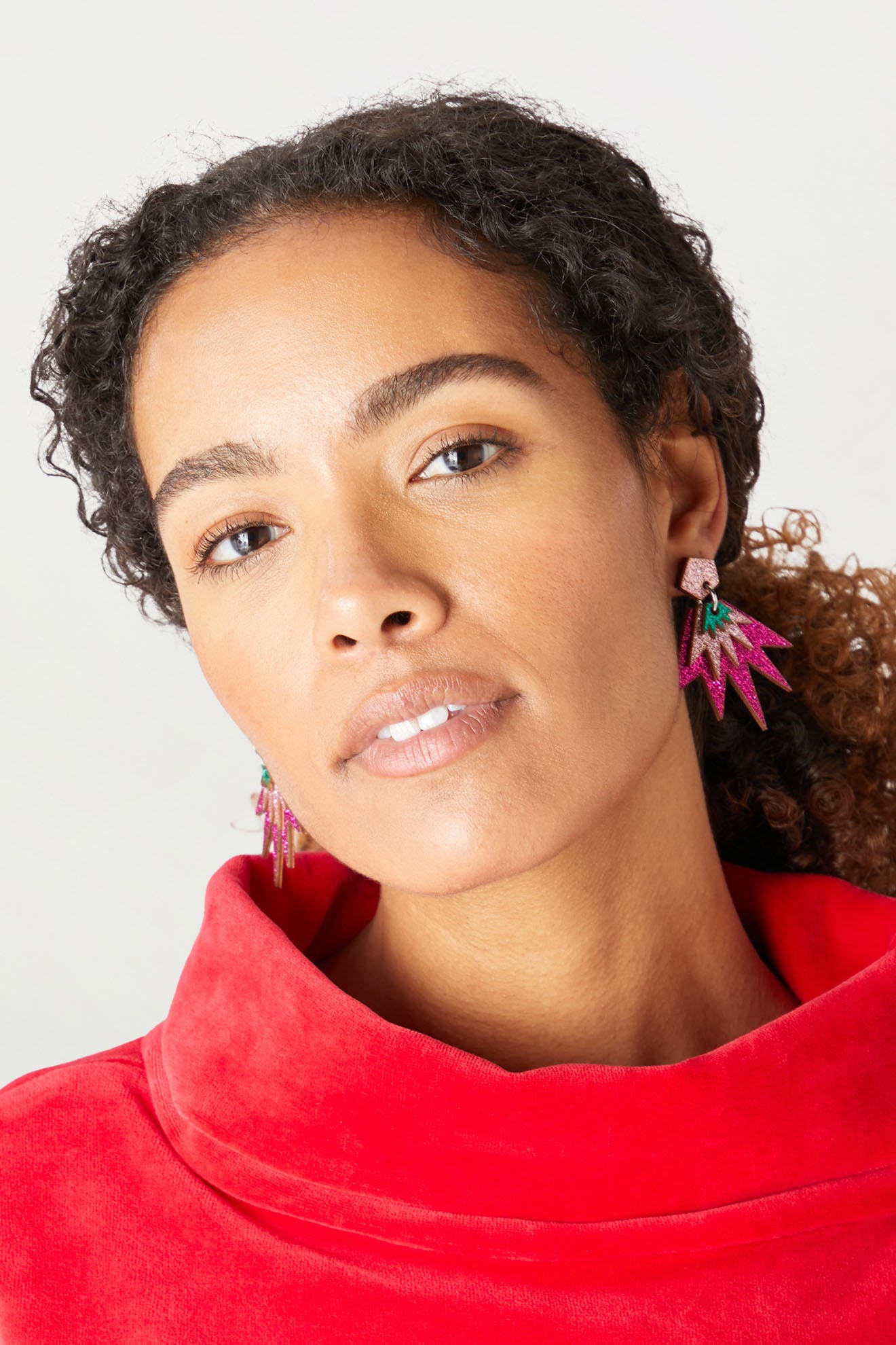 A person with curly hair wearing a red top and the Sparkle Drop Earrings, which are colorful, spiky, and lightweight, looks towards the camera.