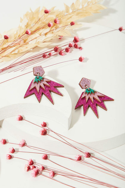 The Sparkle Drop Earrings are pink and gold lightweight geometric drops with green accents, crafted from eco-friendly wood and non-shedding glitter, artfully placed among dried flower stems on a white background.