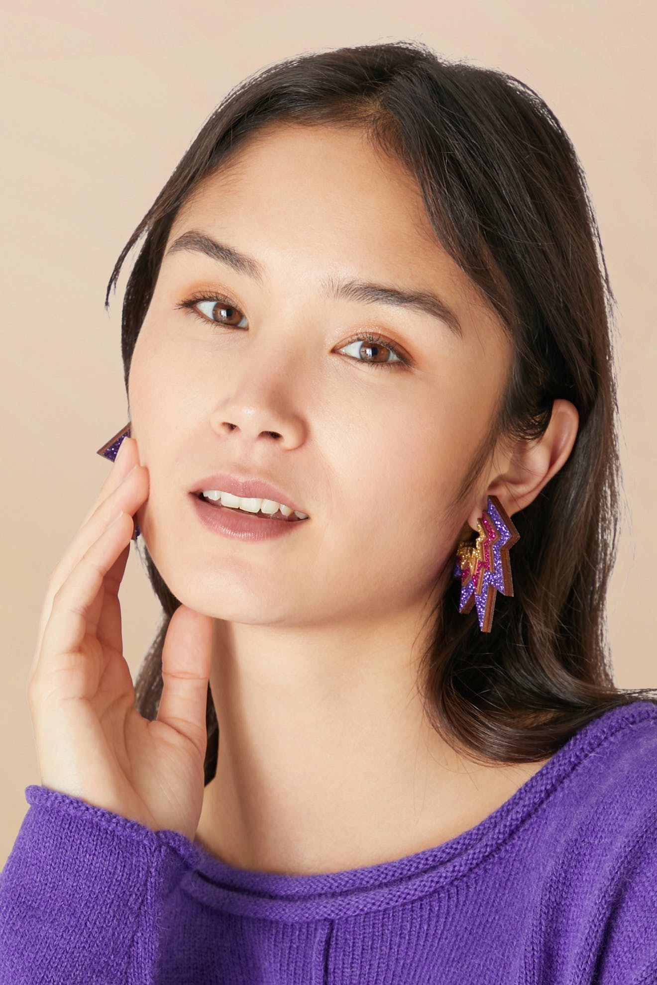 A woman with long dark hair, wearing the vibrant purple Lightning Bolt Sparkle Hoops crafted from eco-friendly wood, paired with a matching purple sweater, touches her cheek with one hand against a light background.