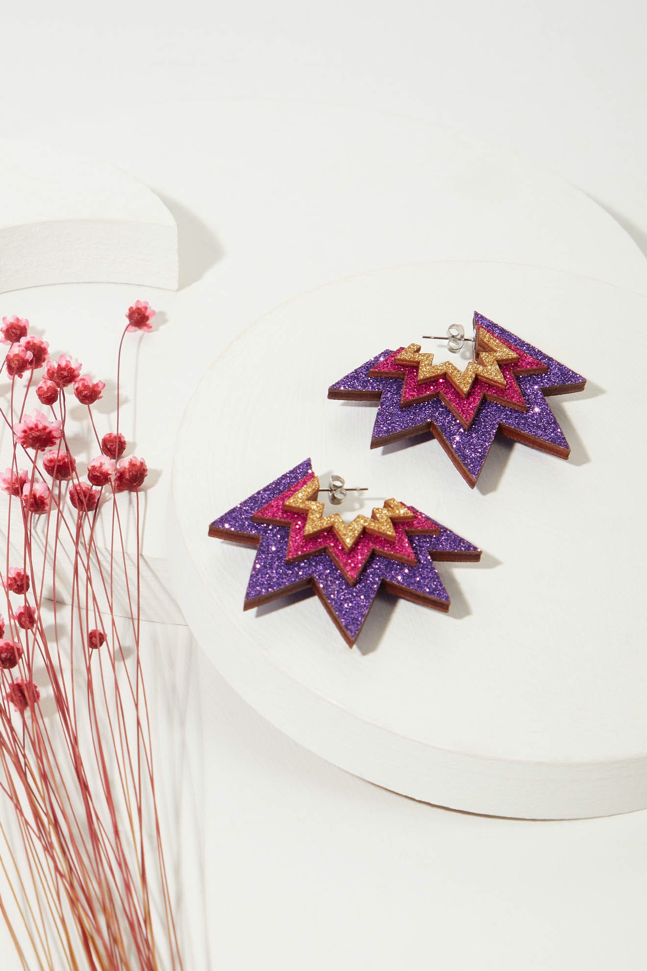 A pair of Laser Cut Sparkle Hoops, geometric and star-shaped, made from eco-friendly wood with purple, pink, and gold glitter, sits on a white circular surface next to a small bouquet of pink dried flowers.