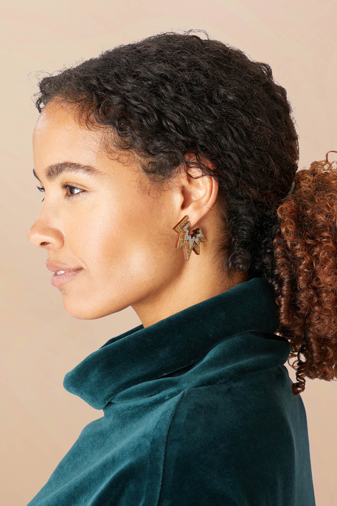 Profile view of a person with curly hair tied back, wearing a teal jacket and gold Mini Laser Cut Sparkle Hoops.