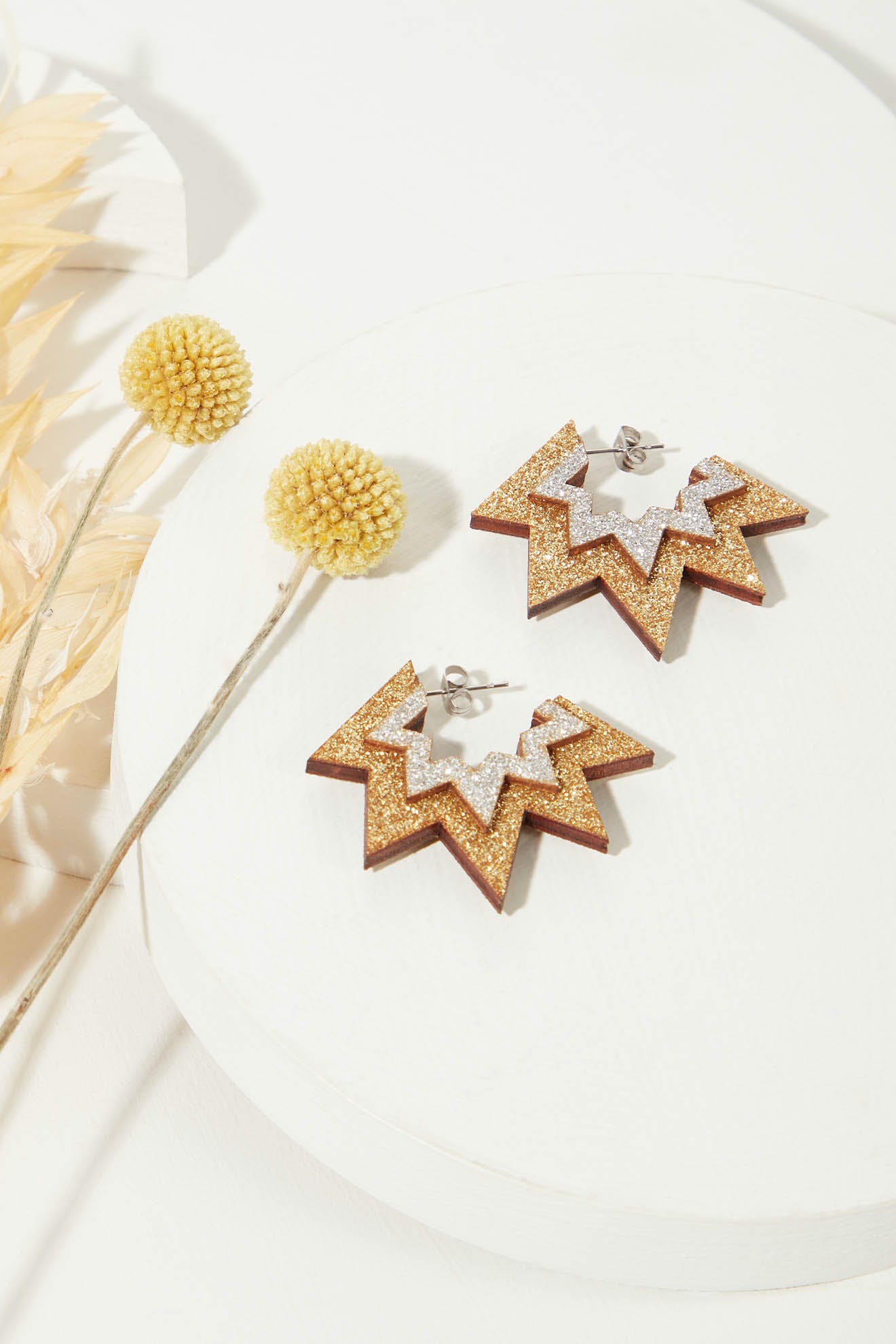 A pair of Mini Laser Cut Sparkle Hoops, featuring star-shaped, lightweight hoops adorned with gold glitter and silver accents on a white circular surface, displayed next to two yellow pompom flowers.