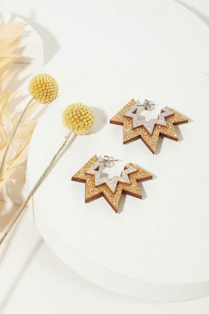 A pair of Mini Laser Cut Sparkle Hoops, featuring star-shaped, lightweight hoops adorned with gold glitter and silver accents on a white circular surface, displayed next to two yellow pompom flowers.