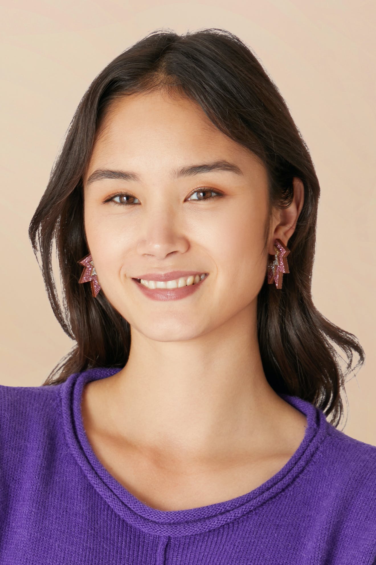 A person with long dark hair is smiling, wearing a purple top and non-returnable Mini Laser Cut Sparkle Hoops in the shape of pink stars.