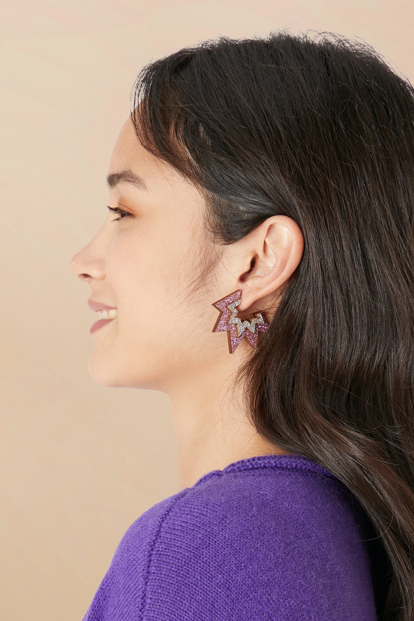 Profile view of a smiling person with long, dark hair wearing a purple sweater and geometric eco-friendly Mini Laser Cut Sparkle Hoops earrings against a beige background.