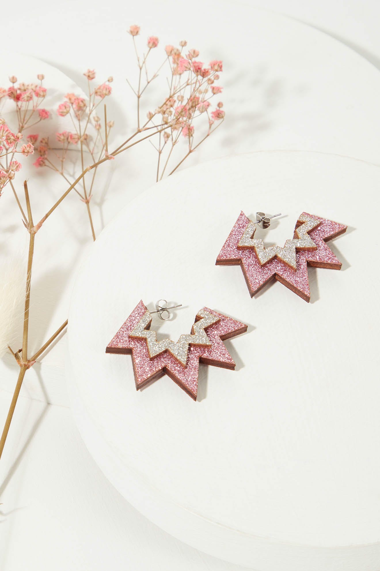 Mini Laser Cut Sparkle Hoops, a pair of star-shaped earrings with pink glitter and gold accents, crafted from eco-friendly wood, are displayed on white circular platforms next to dried pink flowers.