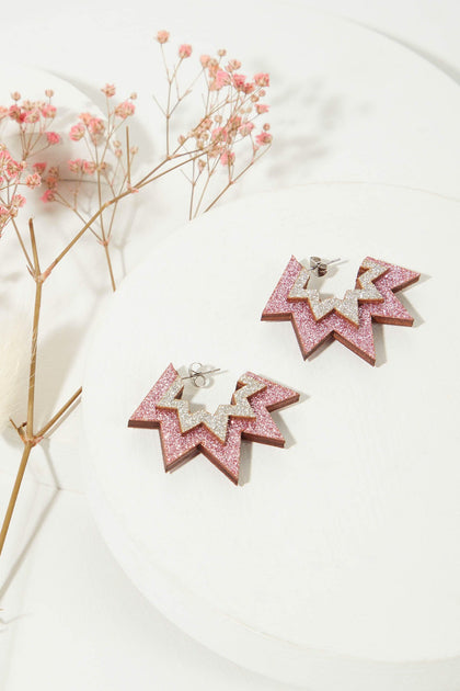 Mini Laser Cut Sparkle Hoops are star-shaped pink and silver glitter earrings displayed on a white circular platform with small pink flowers in the background. These lightweight hoop earrings combine style and comfort effortlessly.