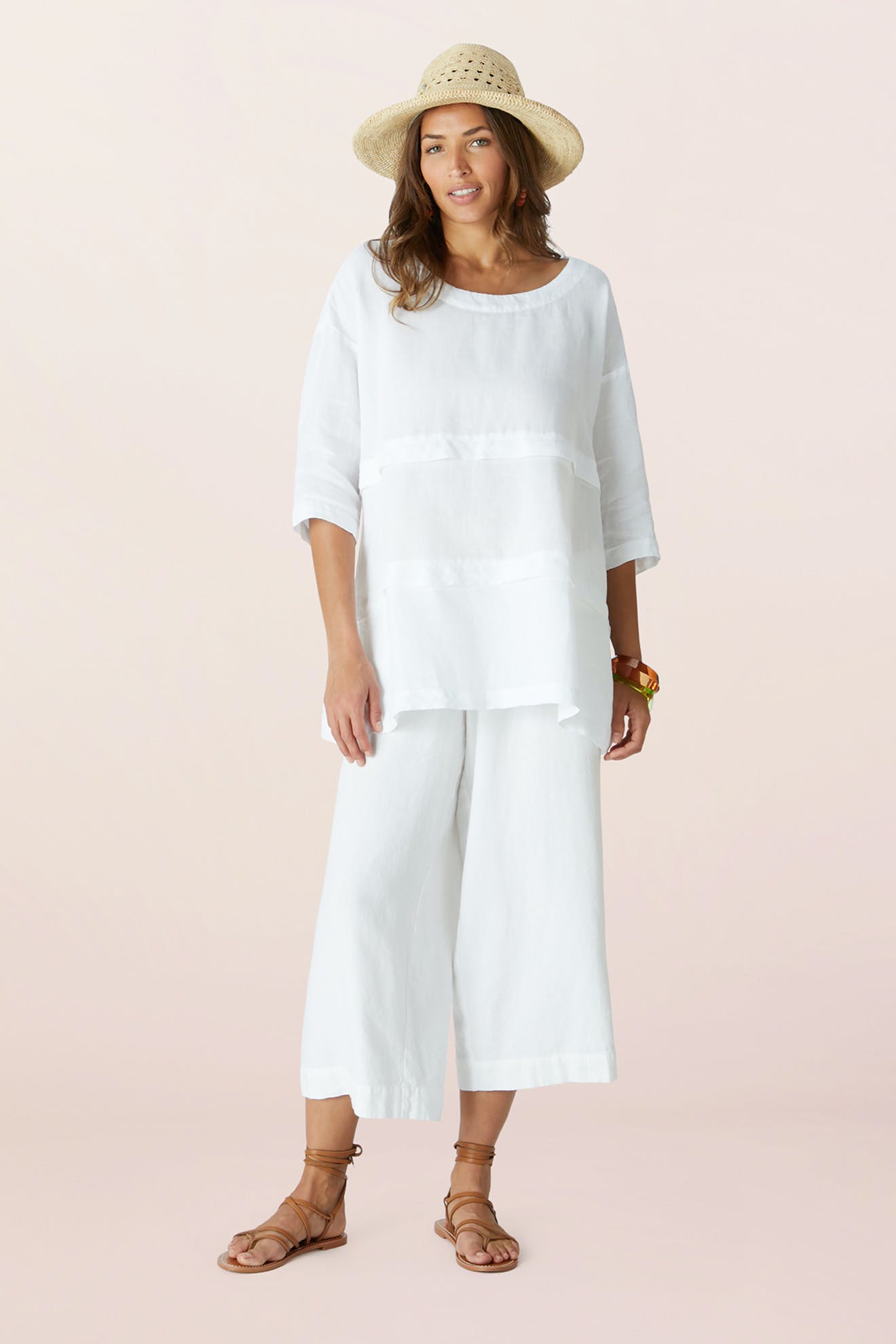A woman stands against a light background wearing the Chapeau Westosia, a close weave hat, paired with a loose white linen top, wide white linen pants, and brown sandals.