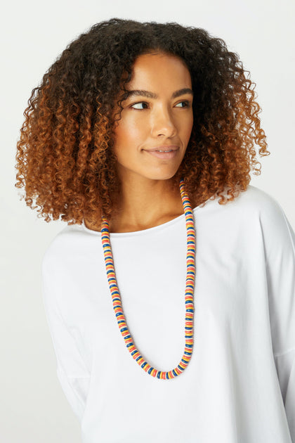 A woman with curly ombré hair and a plain white shirt wears the Mondrian Disc Necklace, featuring multicolored wooden beads, as she looks slightly to the side against a neutral background.