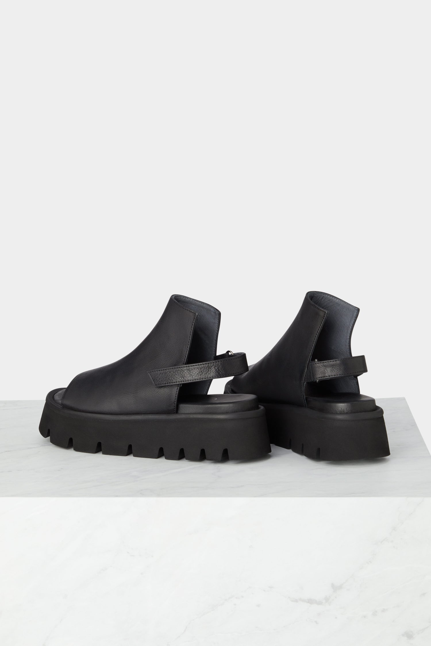 A pair of minimalist-style black leather Buckle Back Sandals on a marble table.