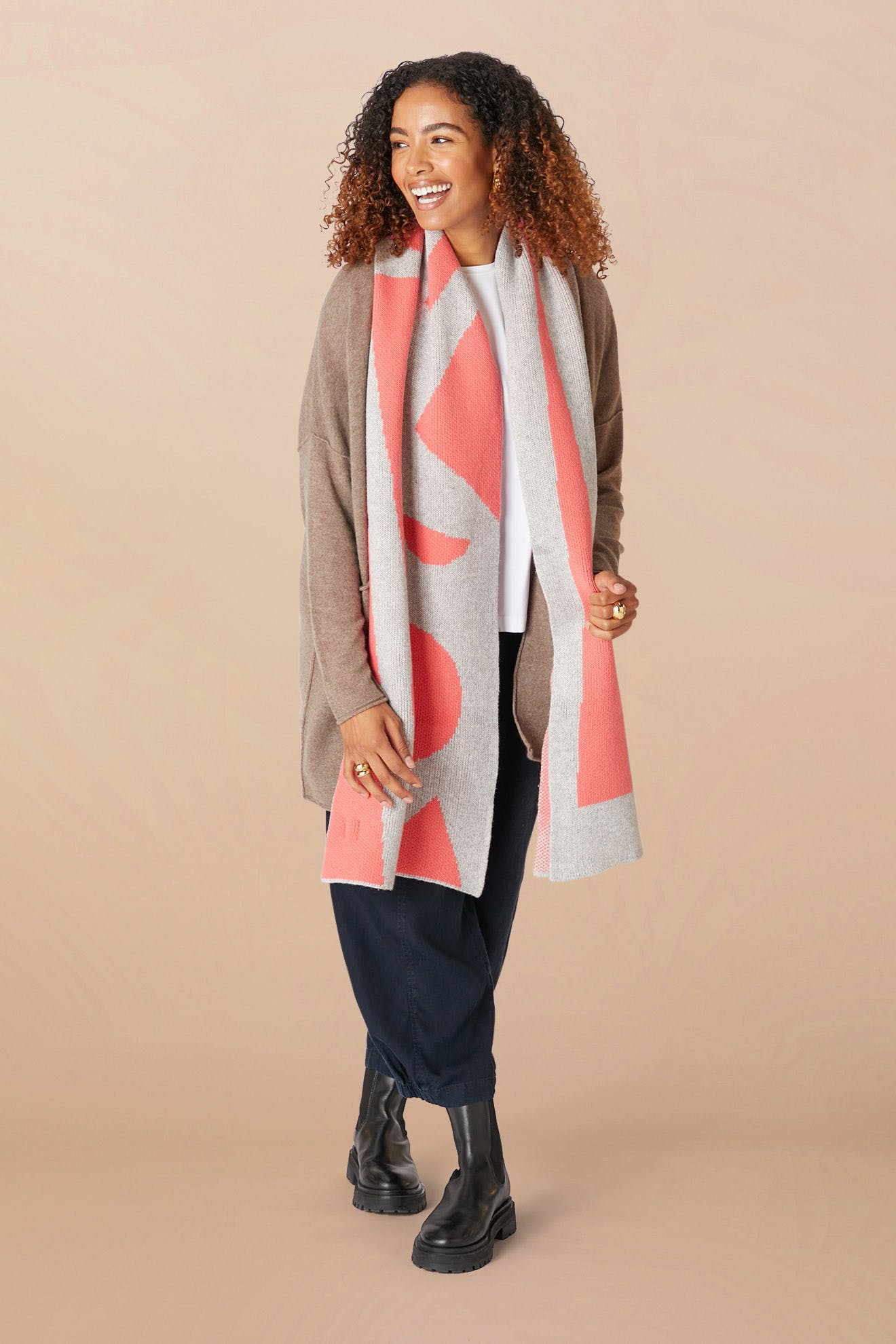 A smiling person with curly hair, wearing a beige cardigan, white shirt, navy pants, black boots, and a Recycled Wool Allsorts Scarf with pink accents made from recycled vegan materials stands against a plain background.