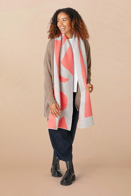 A smiling person with curly hair, wearing a beige cardigan, white shirt, navy pants, black boots, and a Recycled Wool Allsorts Scarf with pink accents made from recycled vegan materials stands against a plain background.