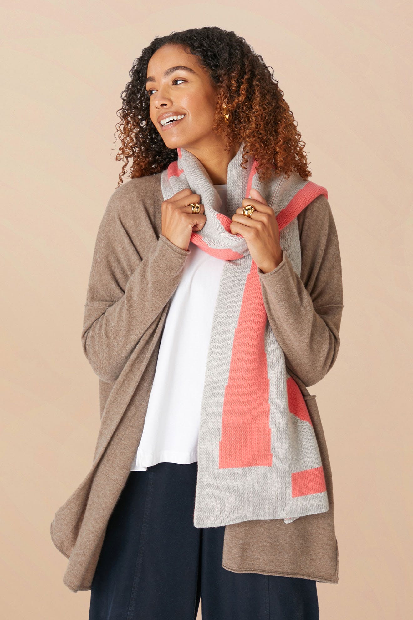 A woman wearing a brown cardigan, white shirt, and the Recycled Wool Allsorts Scarf with pink accents stands against a beige background, showcasing sustainable knitwear crafted from recycled vegan materials.