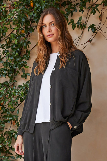 A woman with light brown hair wearing a Fluid Twill Drawstring Jacket and white shirt stands against a wall with climbing plants.