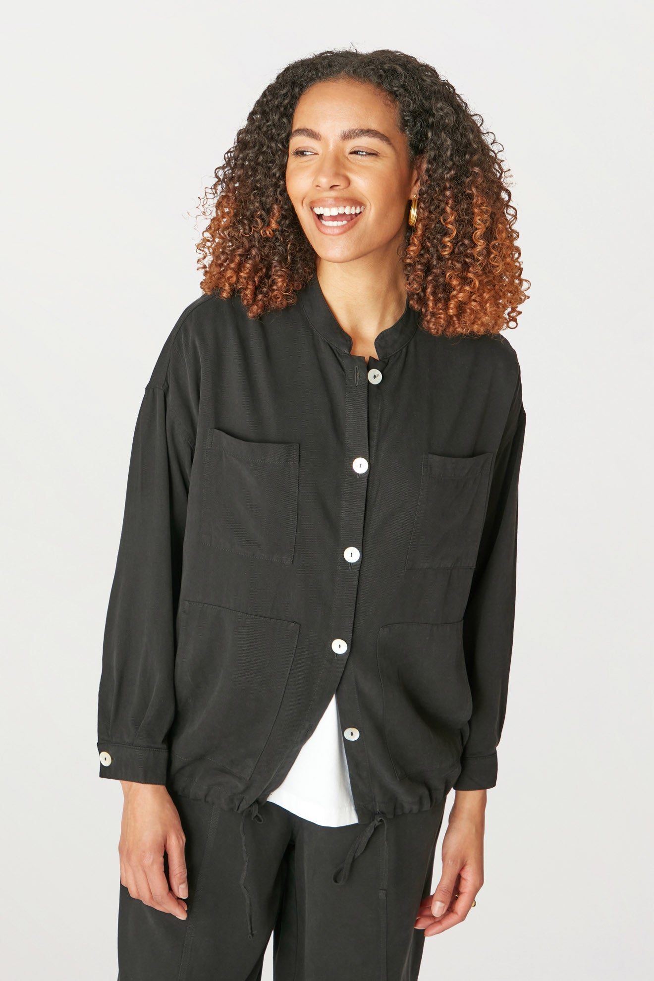 A young woman smiling, wearing a Fluid Twill Drawstring Jacket and white top, standing against a light background.