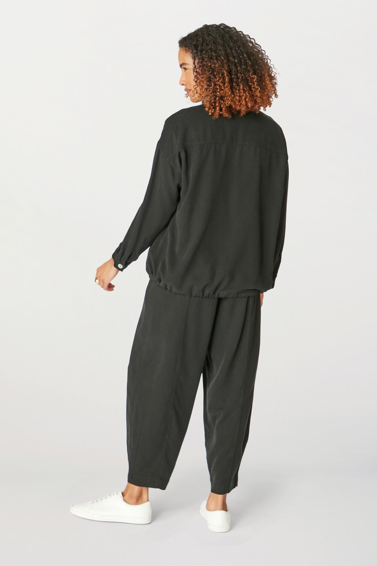 A woman seen from behind, wearing a casual black Fluid Twill Drawstring Jacket and trousers with white sneakers, standing against a light backdrop.