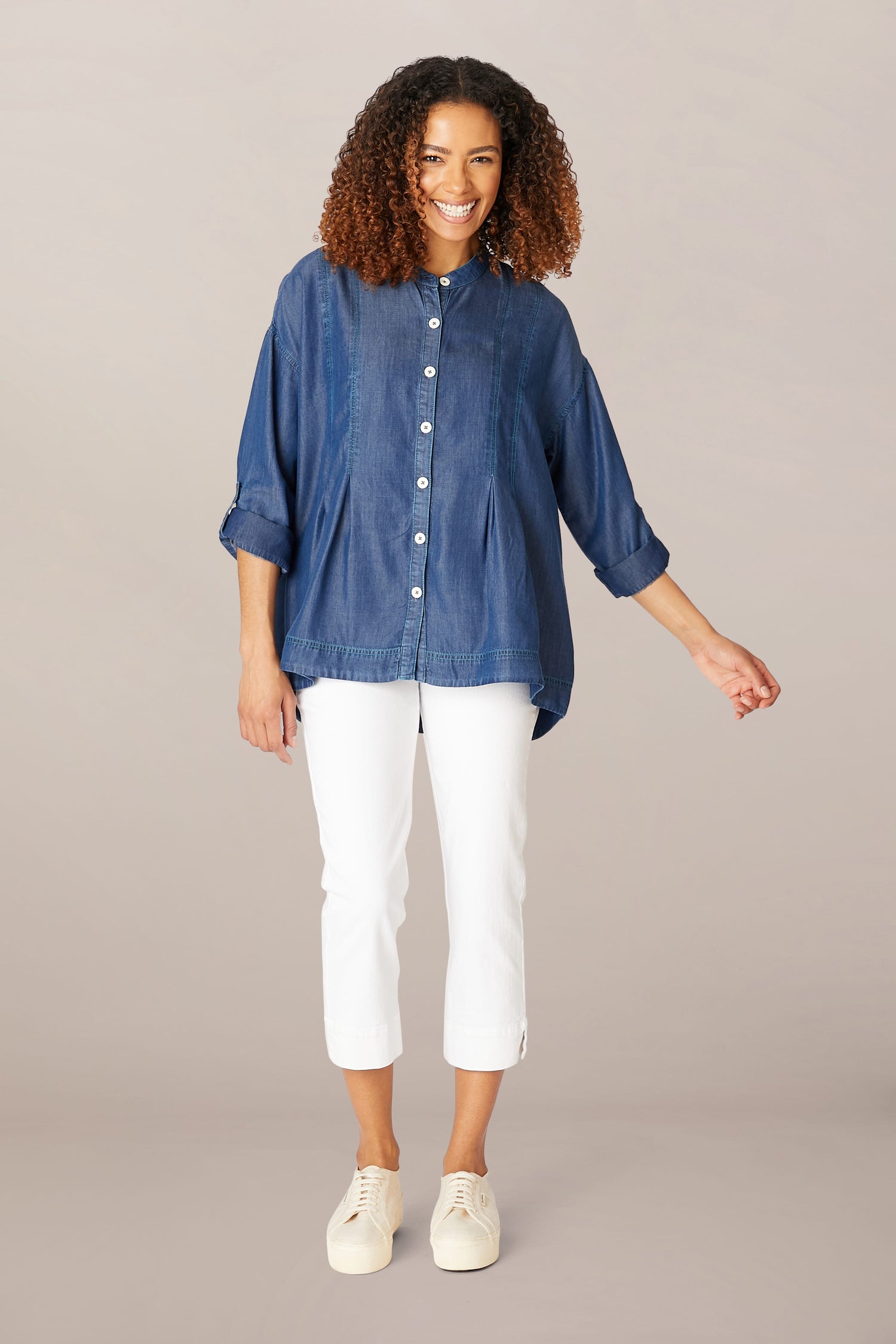 A smiling woman stands posing in a Fluid Denim Relaxed Shirt, white cropped pants, and white sneakers against a neutral background.