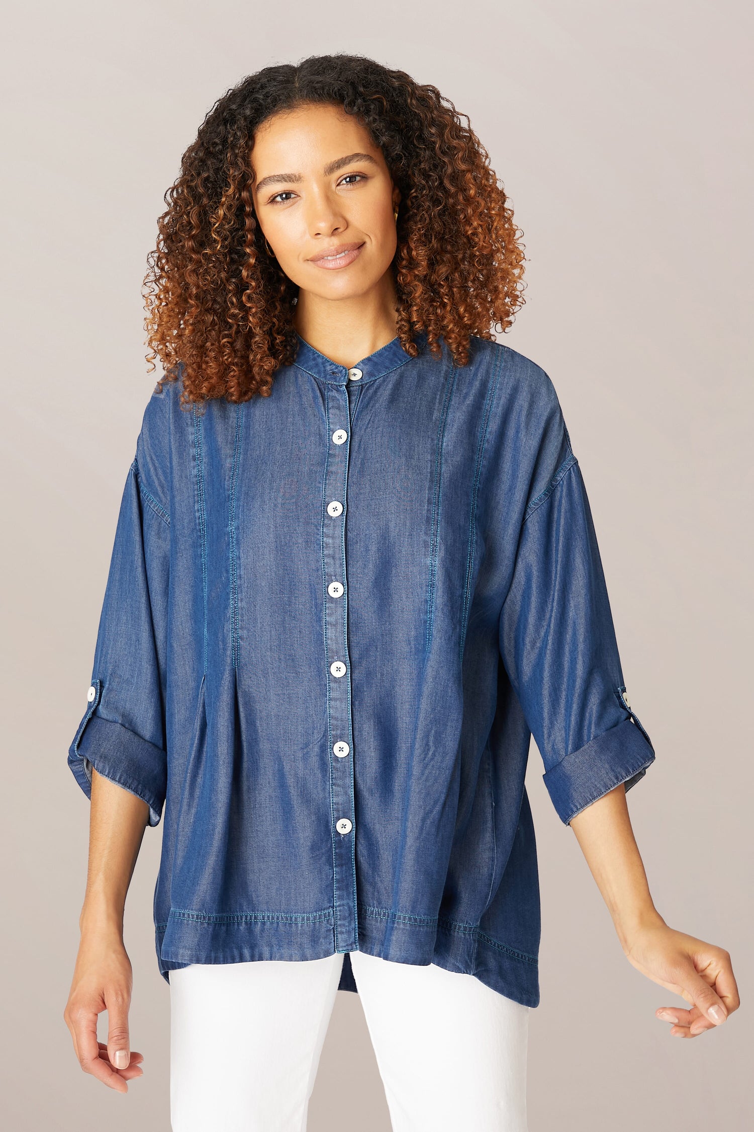 A woman with curly hair, smiling slightly, wearing a Fluid Denim Relaxed Shirt and white pants, standing against a light gray background.